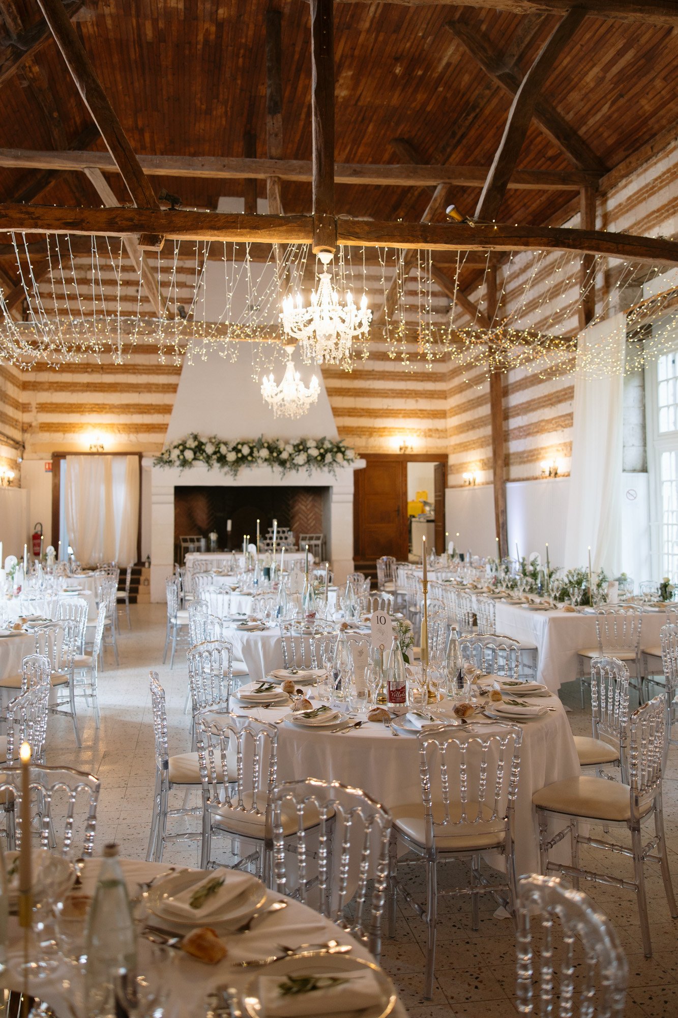 A decorated wedding reception hall with round tables set for guests, clear chairs, floral centerpieces, string lights, and a chandelier hanging from a wooden beam ceiling. Captured by Northern France wedding photographer.