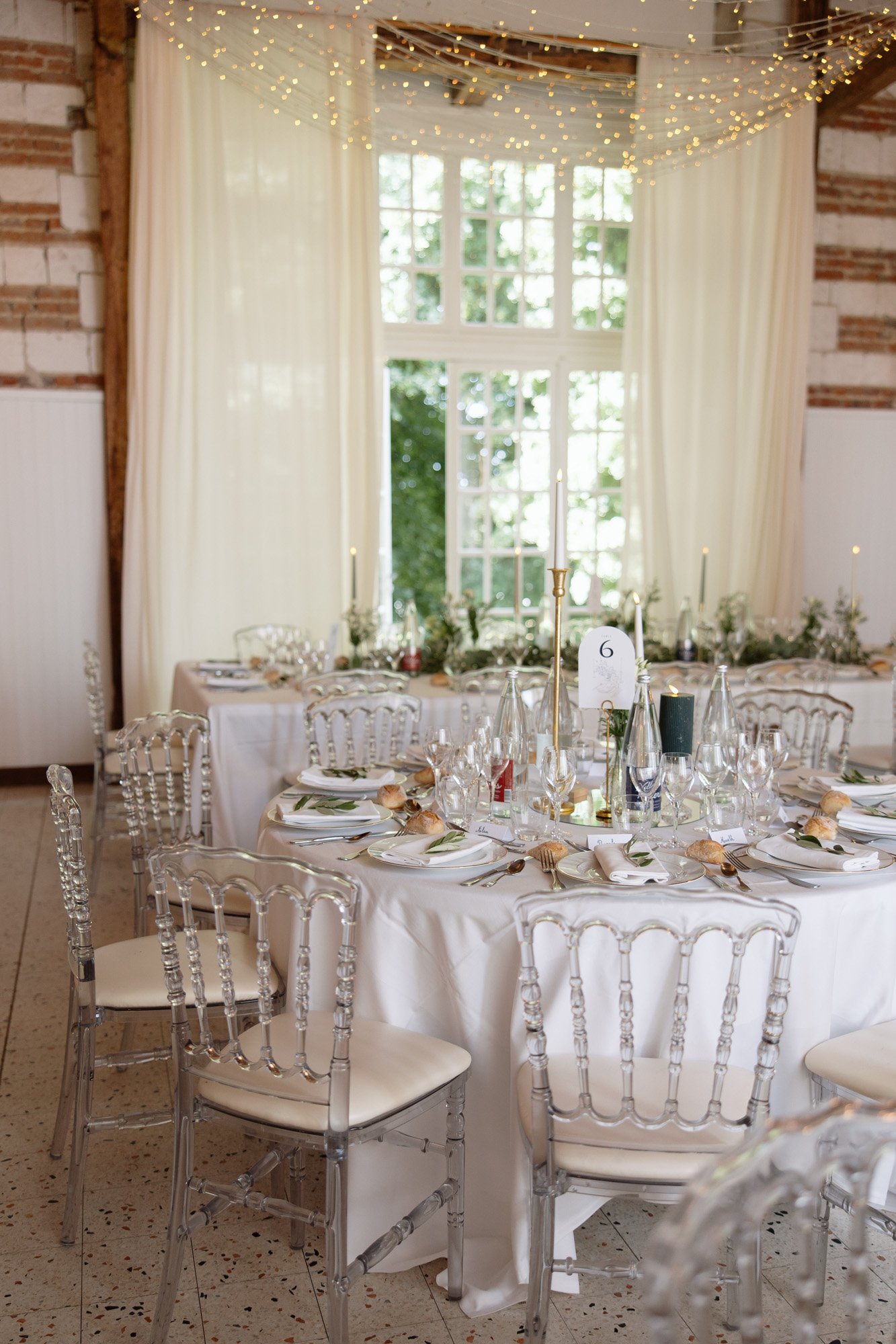 Round banquet table set for a formal event with white linens, elegant glassware, clear chairs, and string lights overhead in a bright, airy room with large windows and curtains. Captured by Northern France wedding photographer.