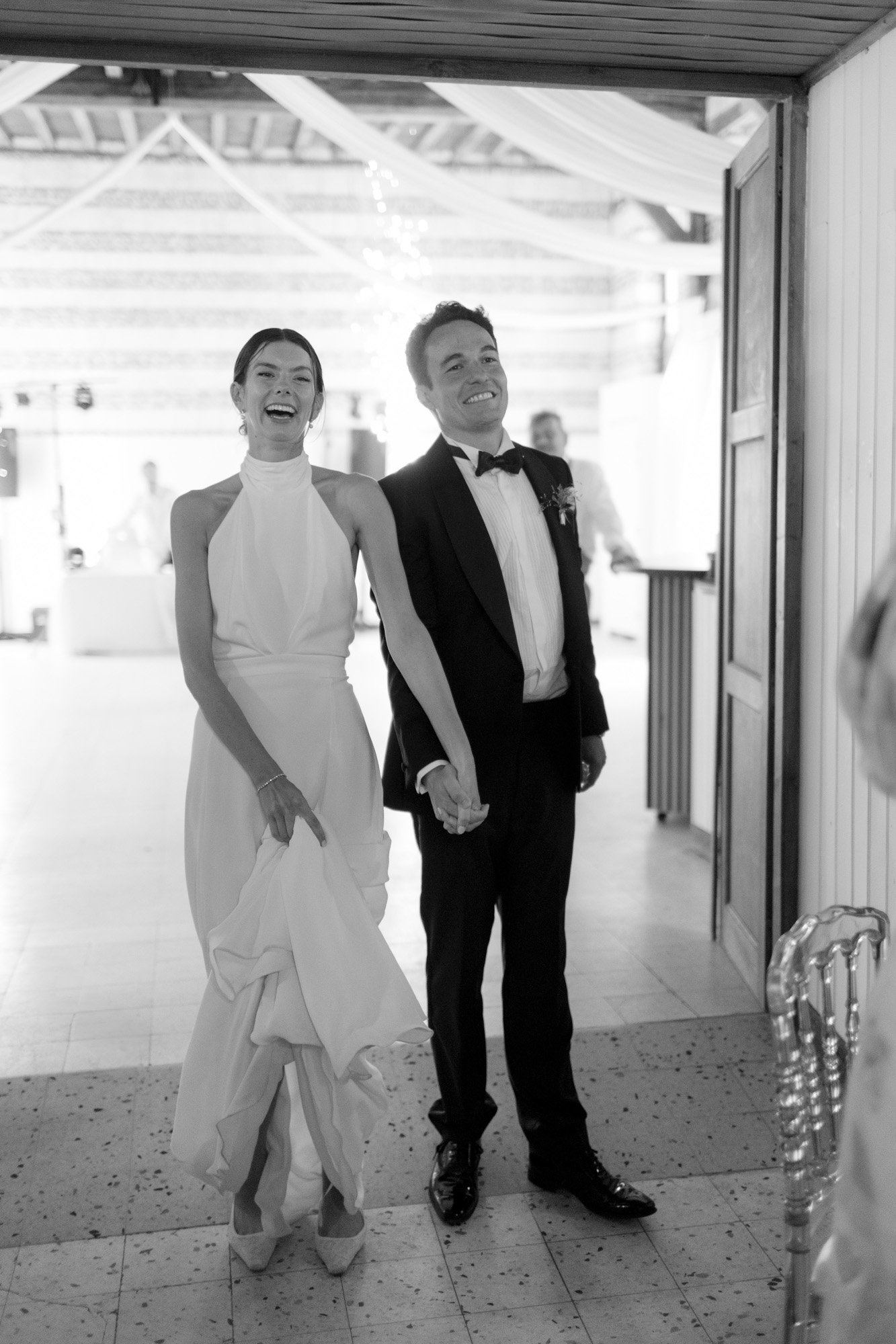 A bride and groom, holding hands and smiling, enter a decorated indoor venue, dressed in formal wedding attire. Captured by Northern France wedding photographer.