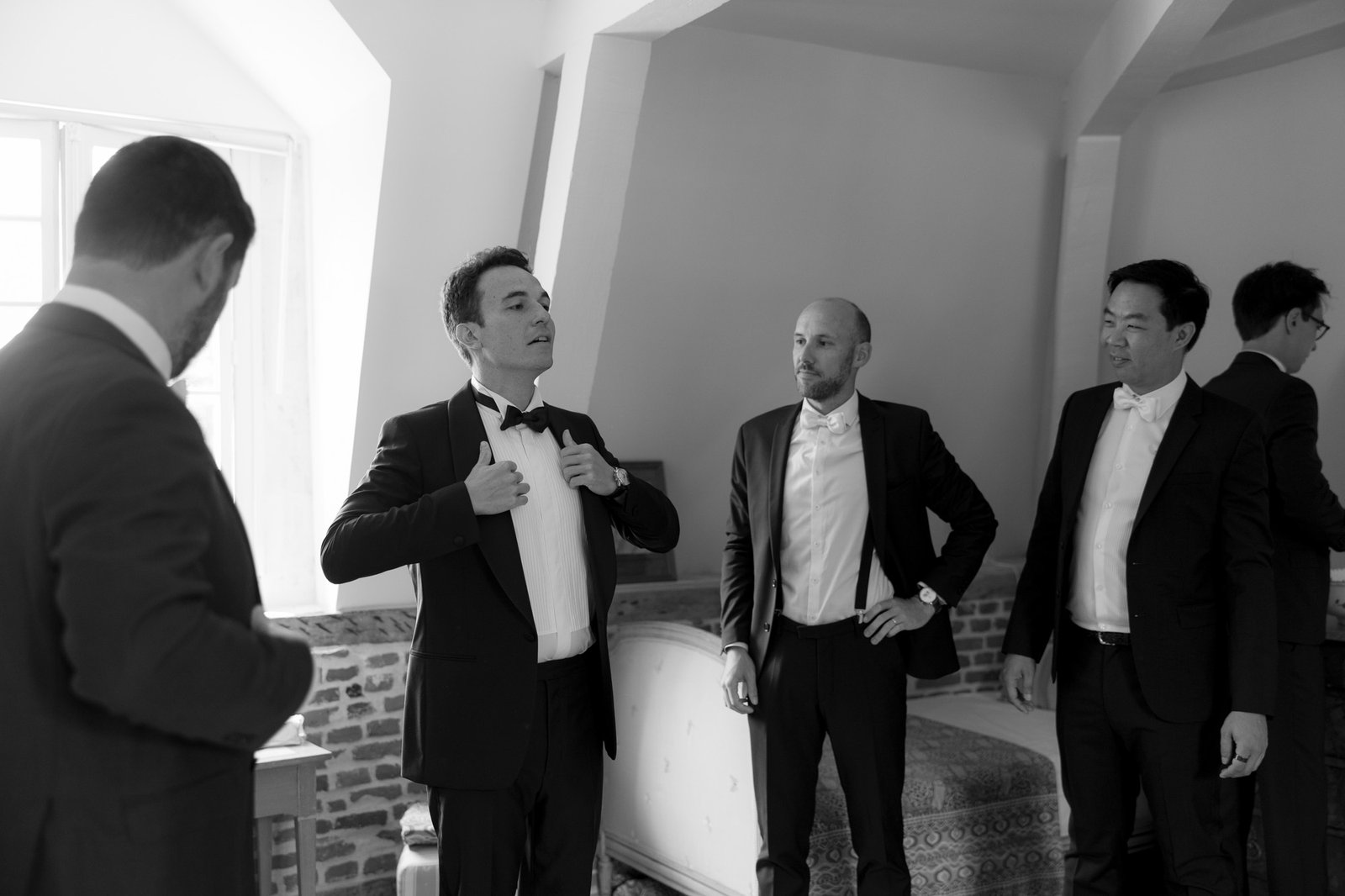 Four men in tuxedos stand in a room with light streaming through a window; one man adjusts his bow tie while the others watch.