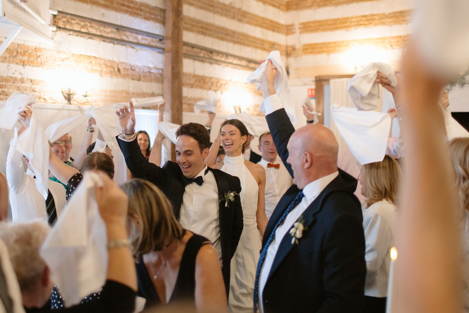 Wedding guests and a bride and groom wave napkins in the air while celebrating at a reception in a rustic indoor venue with wooden beams and white walls.