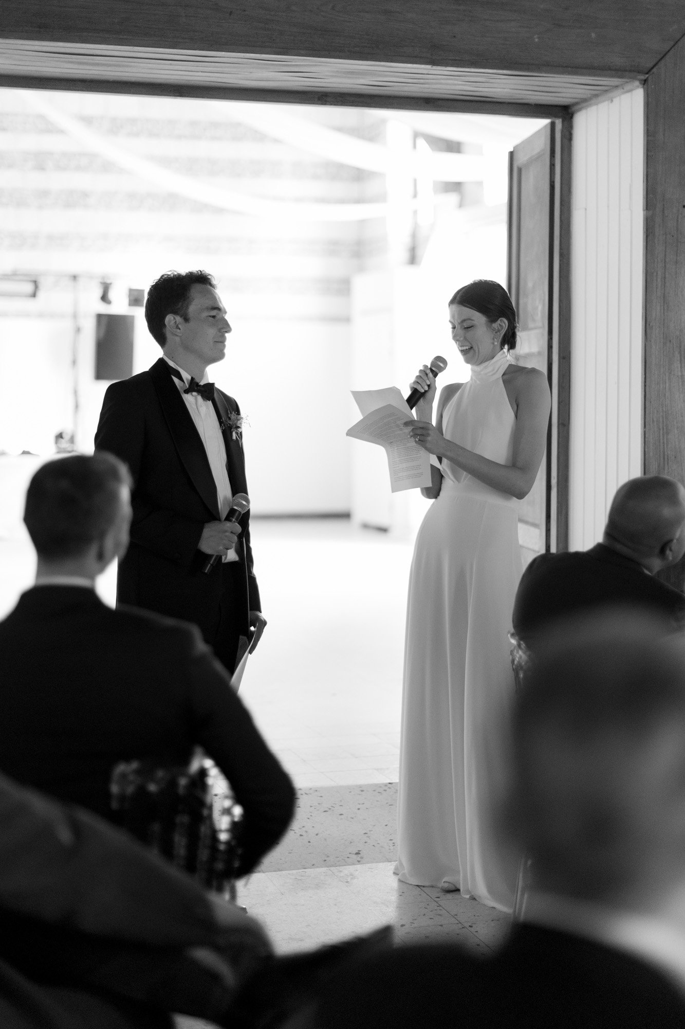 A woman in a white dress reads from a paper while holding a microphone beside a man in a tuxedo; seated guests listen in the foreground.