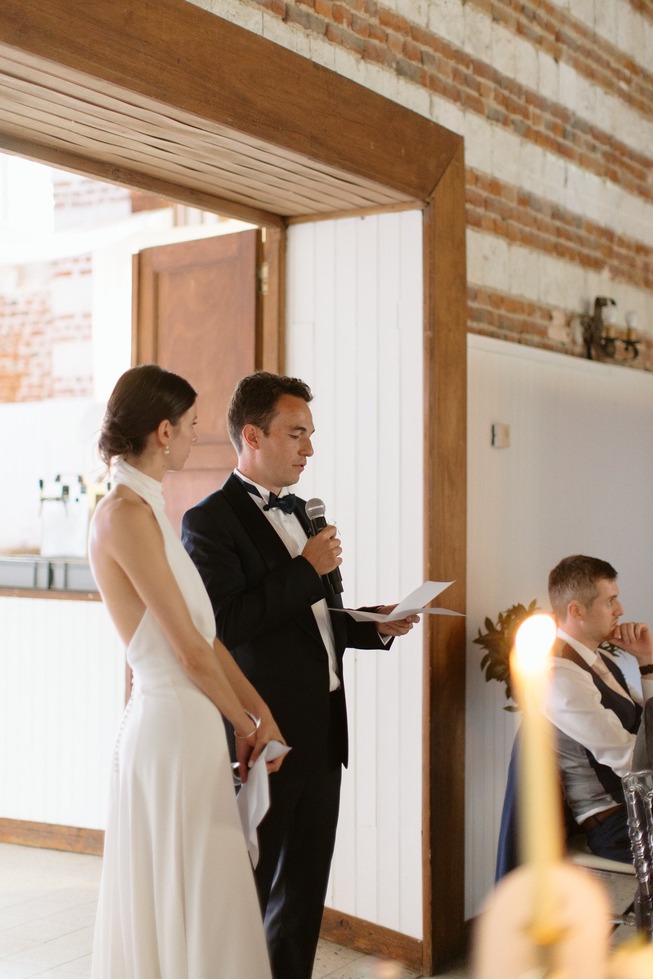A man in a tuxedo speaks into a microphone while holding a paper, standing beside a woman in a white dress at an indoor event. Another person sits nearby.
