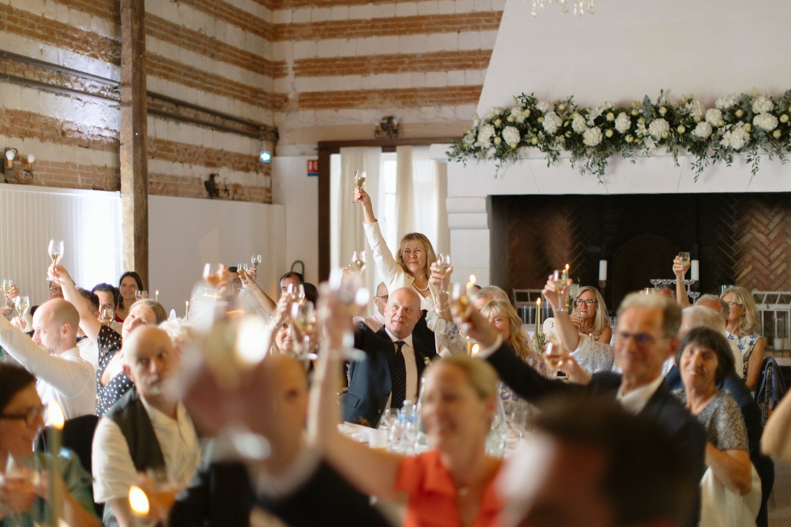 A group of people seated at tables indoors raise glasses in a toast at a formal event, with floral decorations on the fireplace in the background.