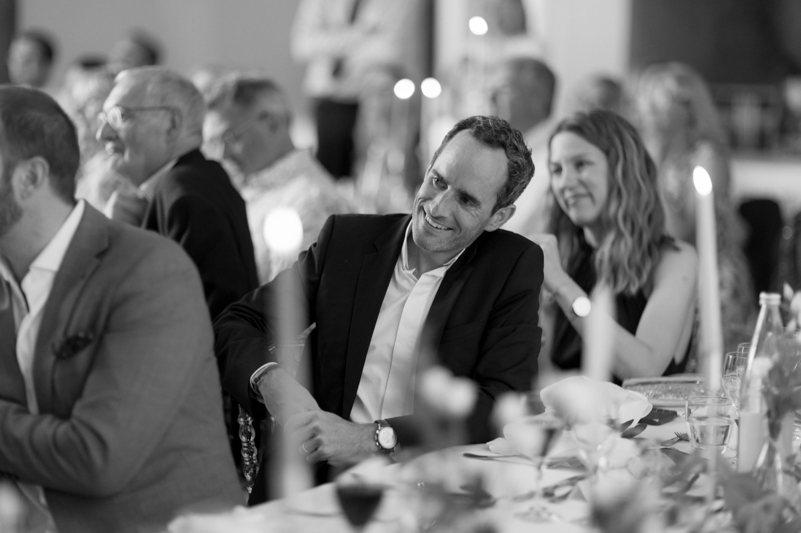 A man in a suit sits at a table, smiling and listening, surrounded by other formally dressed people at a social event.