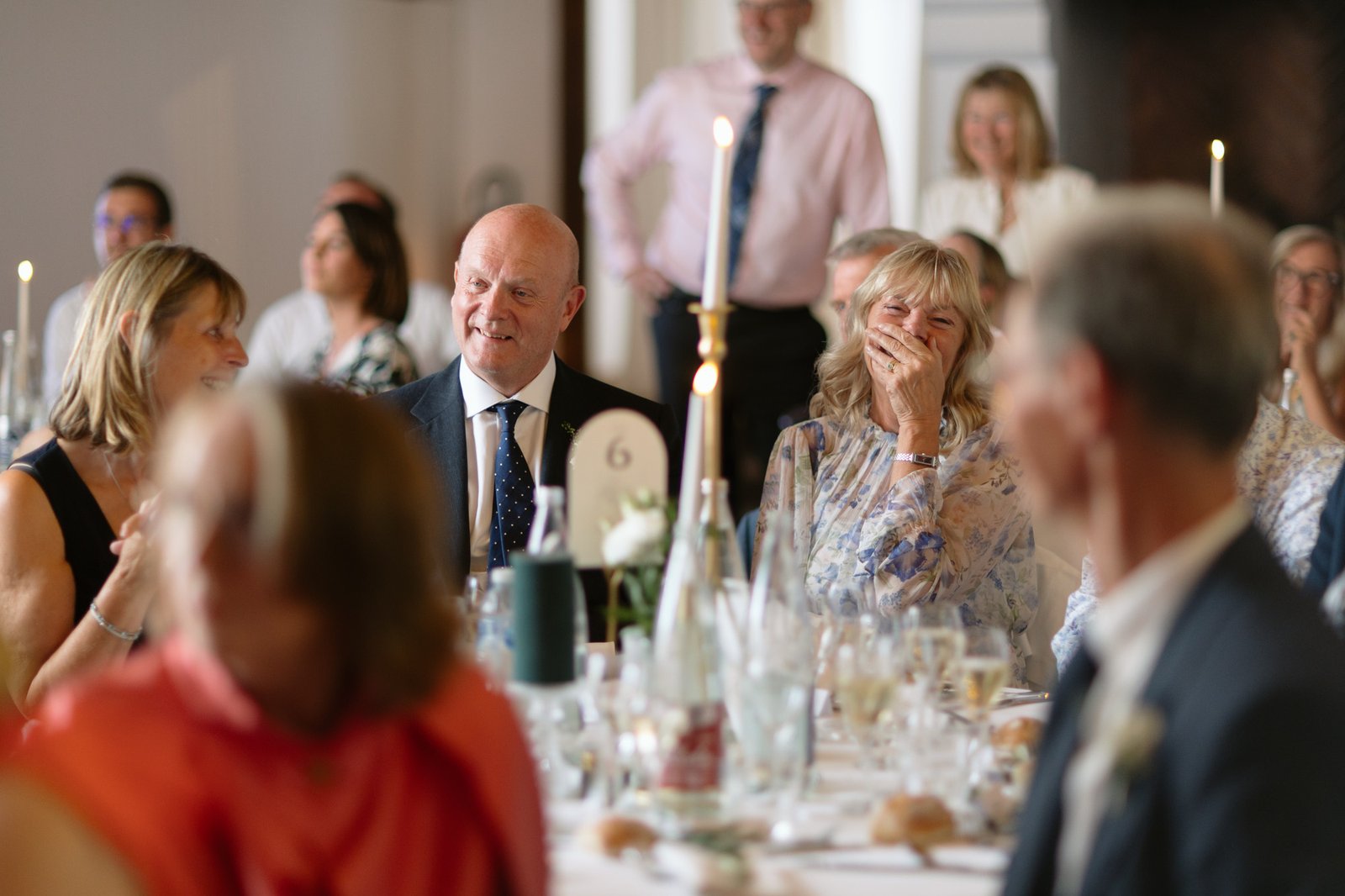 People seated at a formal event table, dressed in semi-formal attire, smiling and laughing, with candles and drinks on the table.