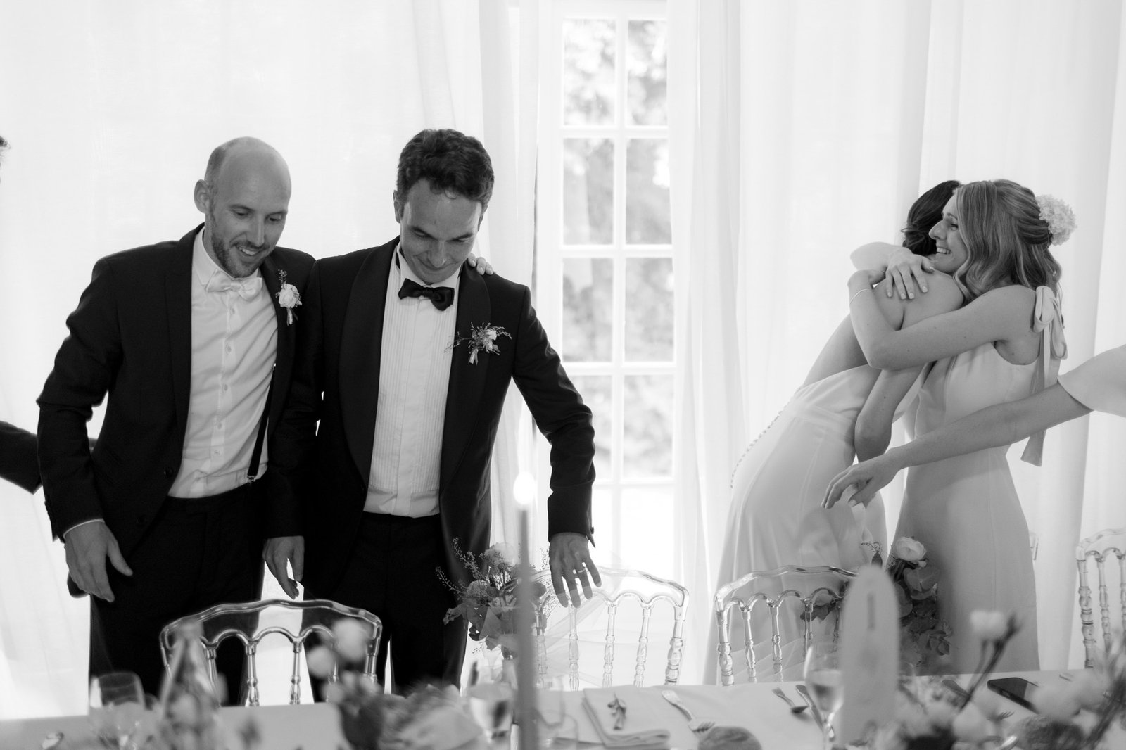 Four people at a formal event; two men in suits are standing by chairs, while two women in dresses embrace near a table with decorations.