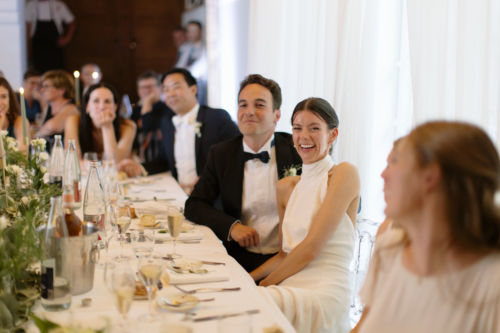 A bride and groom sit together at a table, smiling and surrounded by guests during a wedding reception. Captured by Northern France wedding photographer.