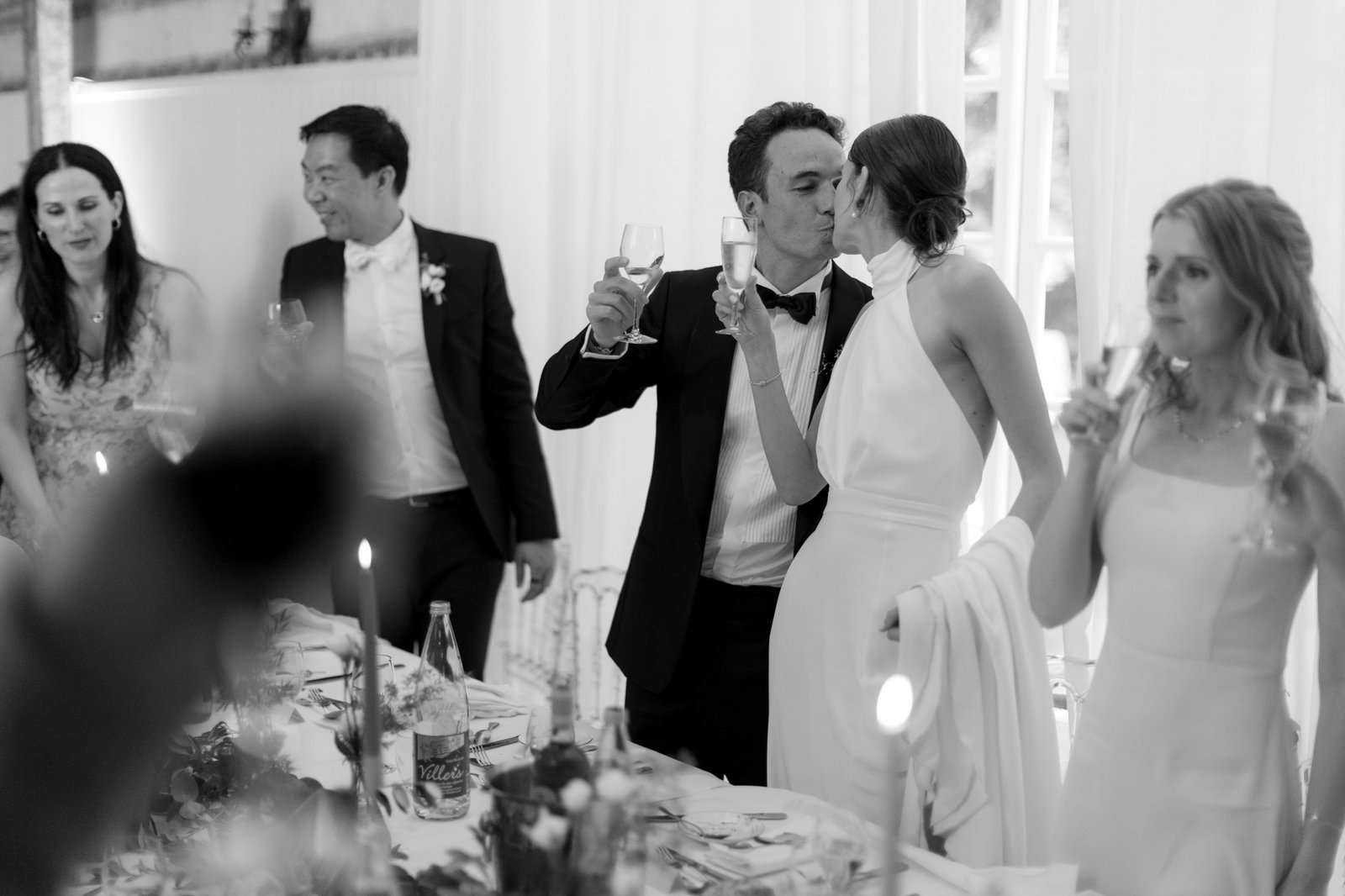 A bride and groom kiss and raise glasses at a wedding reception table, surrounded by guests holding drinks; the scene is in black and white.