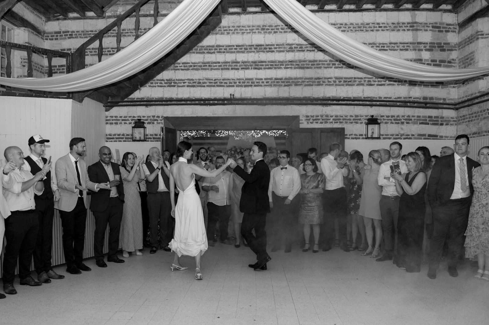 A couple dances in the center of a room, surrounded by guests watching and clapping at what appears to be a wedding reception. The scene is indoors with draped fabric overhead.