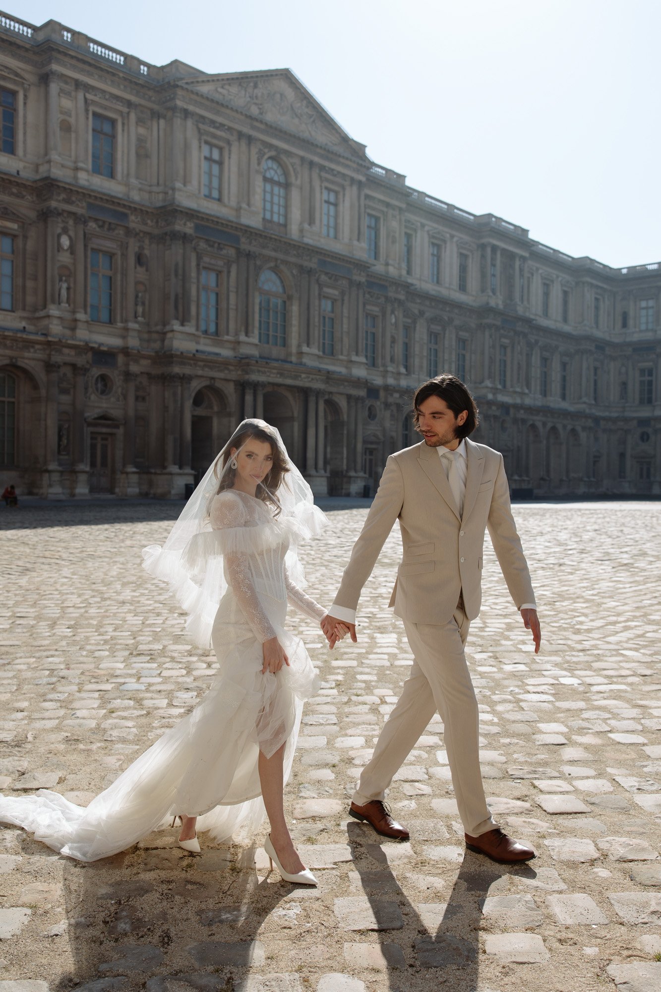 A bride in a white gown and veil walks hand in hand with a groom in a light suit across a cobblestone courtyard in front of a historic stone building. Paris city elopement photographer.