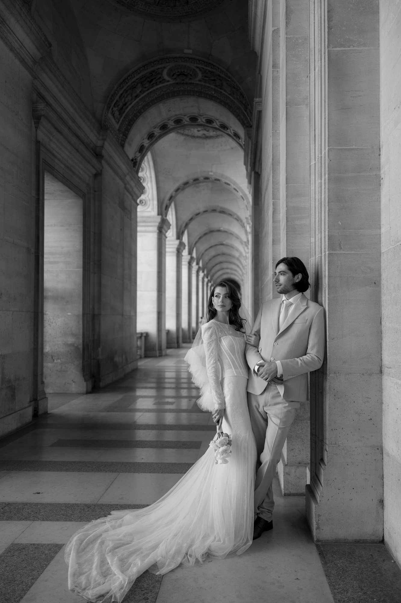 A bride in a long gown and a groom in a suit stand together in an arched corridor, with the bride holding a bouquet and looking to the side.