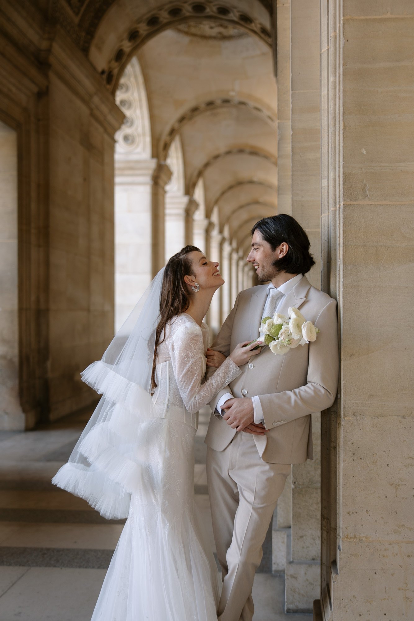 Bride in a white gown and veil smiles at groom in a beige suit holding white flowers; they stand in an arched stone hallway. Paris city elopement photographer.