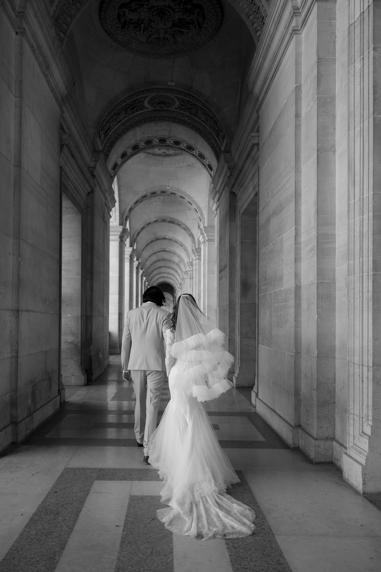 A bride and groom walk together down an ornate, arched hallway with stone walls and checkered floors, photographed from behind in black and white.