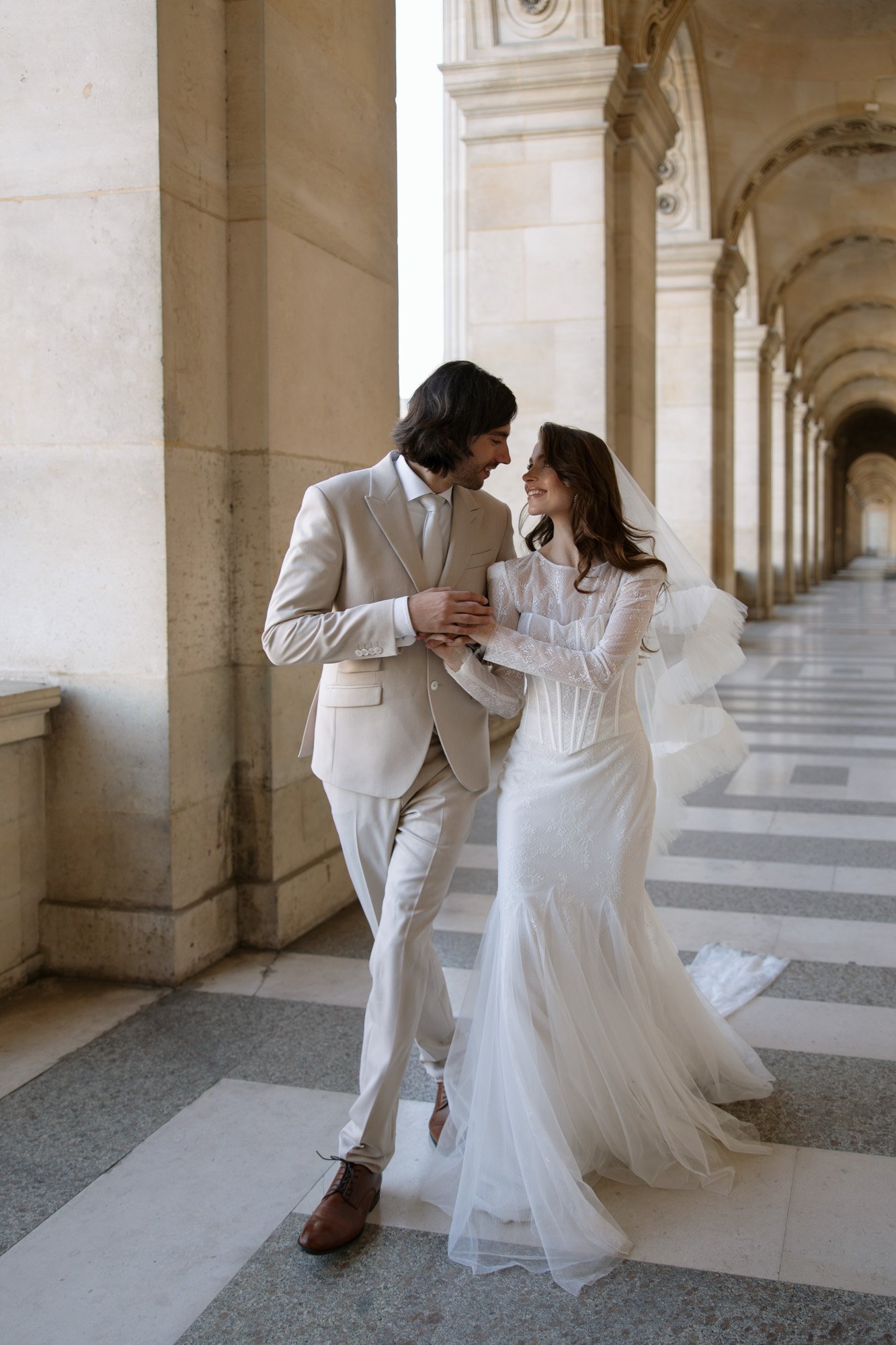 A bride and groom in formal wedding attire walk together under stone arches, smiling and holding hands. Paris city elopement photographer.