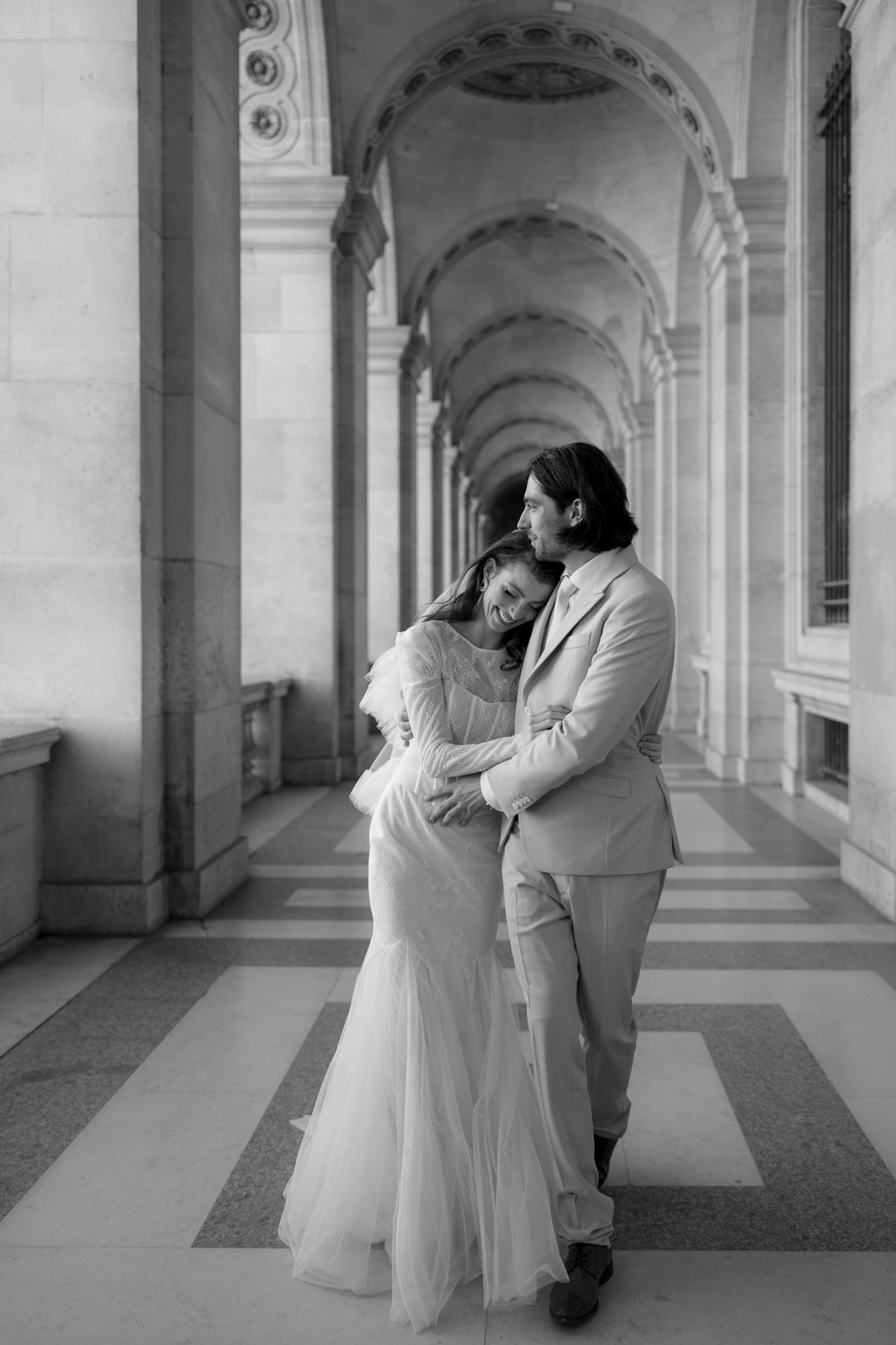 A couple in formal attire embraces under an ornate, arched hallway. The woman in a wedding dress leans on the man, who is in a suit, as he kisses her forehead. Captured by France wedding photographer.