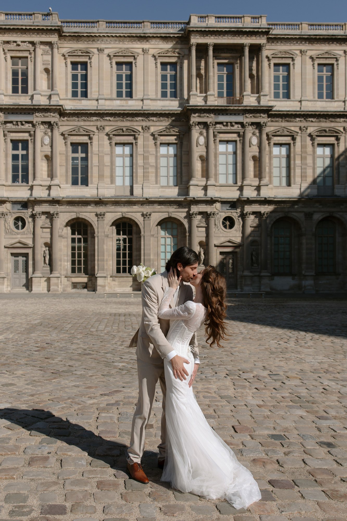 A bride and groom kiss outdoors in front of a historic stone building with large windows and columns. Paris city elopement photographer.