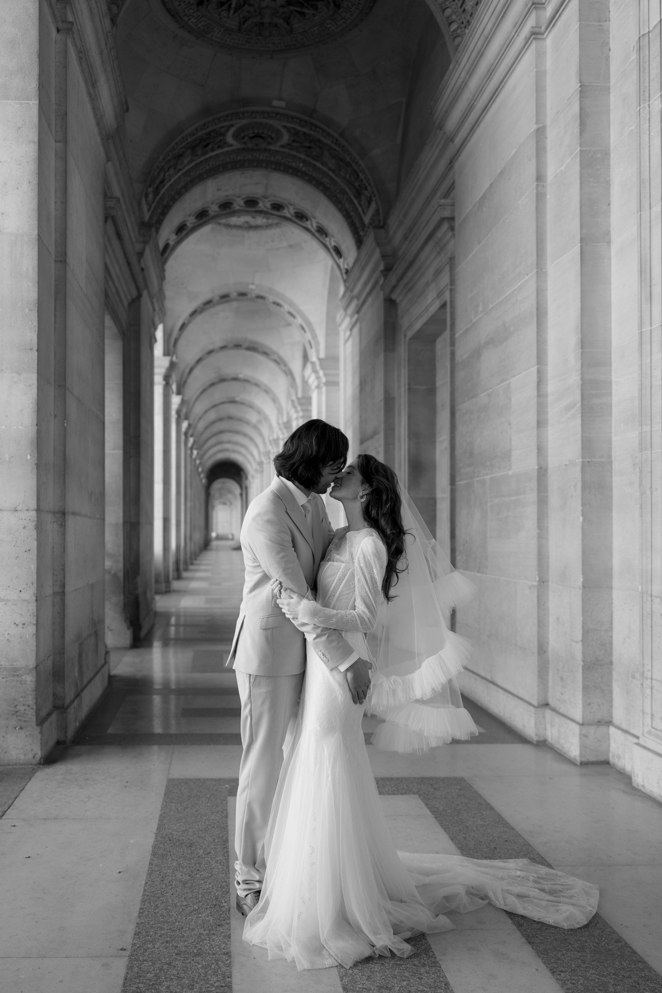 A bride and groom kiss under an ornate stone archway in a grand hallway, both dressed in formal wedding attire. Paris city elopement photographer.