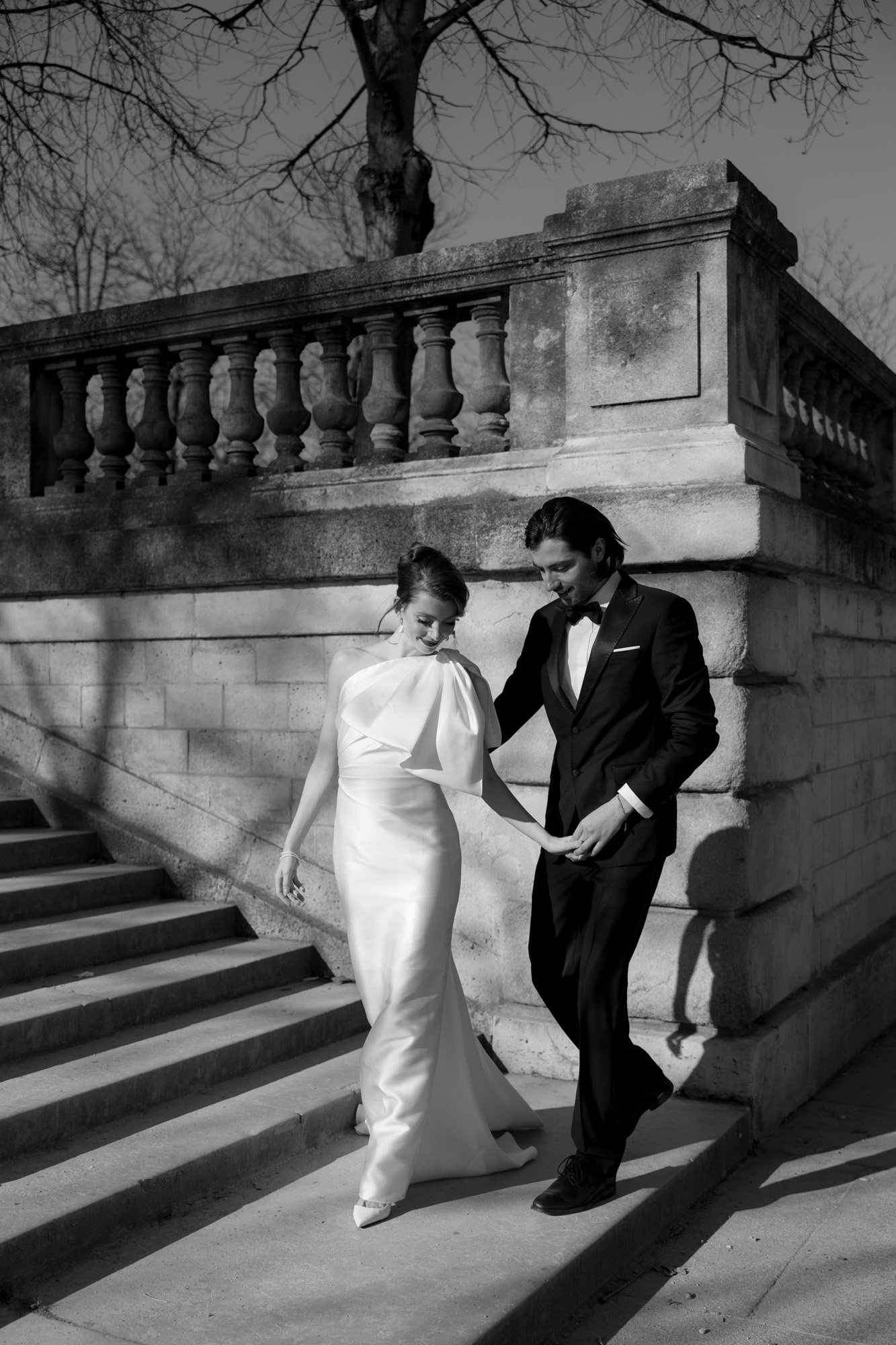 A bride in a white dress and a groom in a suit walk down stone steps outdoors, holding hands, beside a balustrade and bare tree branches.