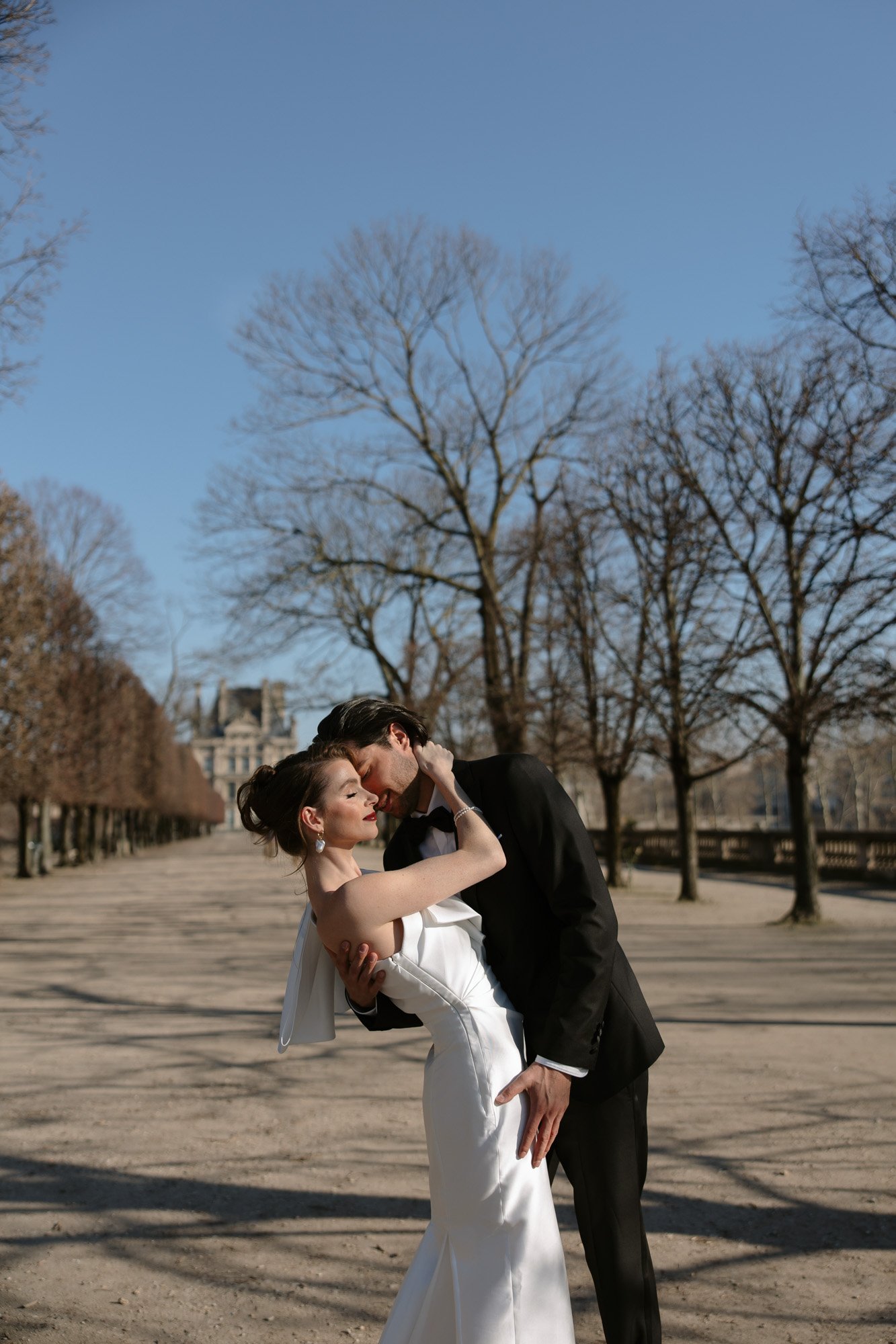 A bride and groom in formal attire embrace and pose on a tree-lined path in a park under a clear blue sky. Paris city elopement photographer.