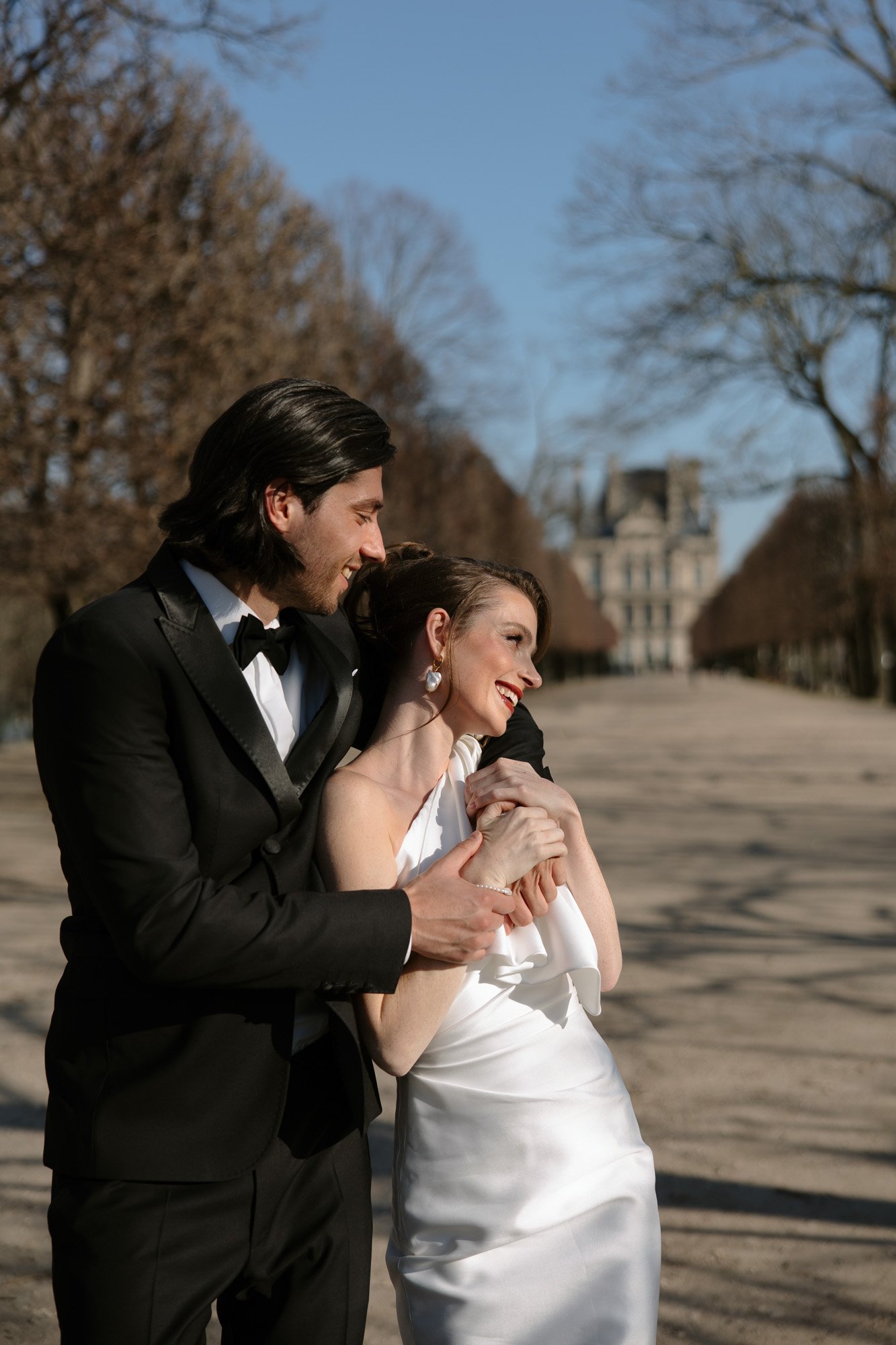 A couple dressed in formal attire, with the woman in a white dress and the man in a black suit, stand outside on a tree-lined path on a sunny day.