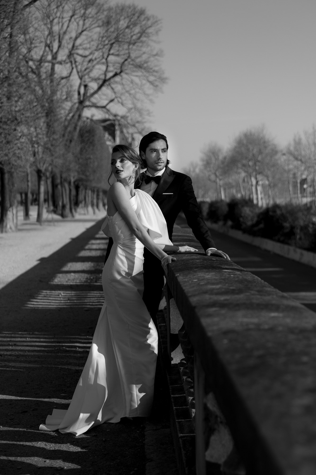 A couple dressed in formal wedding attire stands by a stone railing in an outdoor setting with bare trees lining a pathway. Paris city elopement photographer.