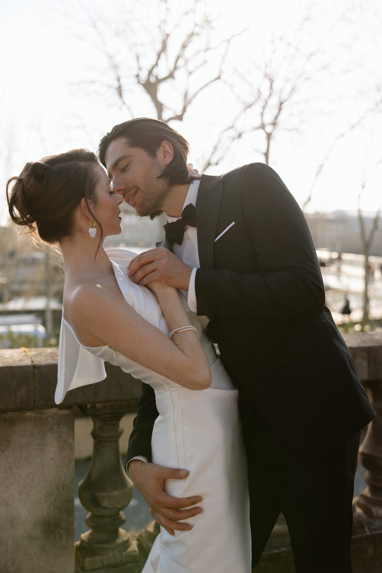 A bride and groom in formal attire share a romantic moment outdoors, with the groom leaning in to kiss the bride by a stone balustrade. Paris city elopement photographer.