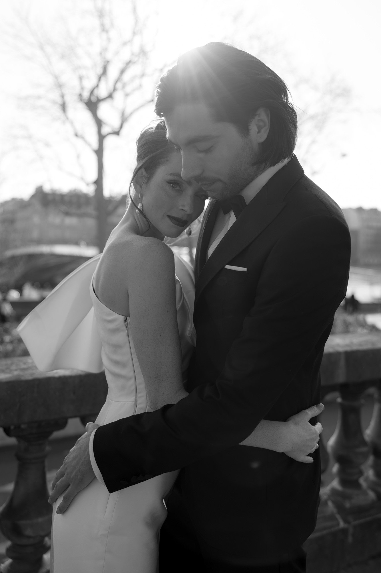 A couple dressed formally embrace outdoors in sunlight, with bare trees and a blurred cityscape in the background.