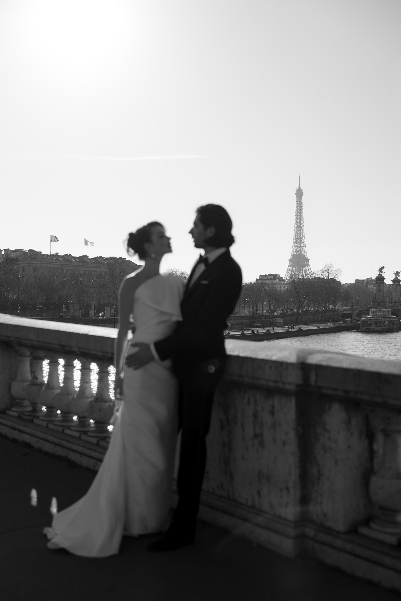 A bride and groom stand together on a bridge in Paris with the Eiffel Tower visible in the background on a sunny day.