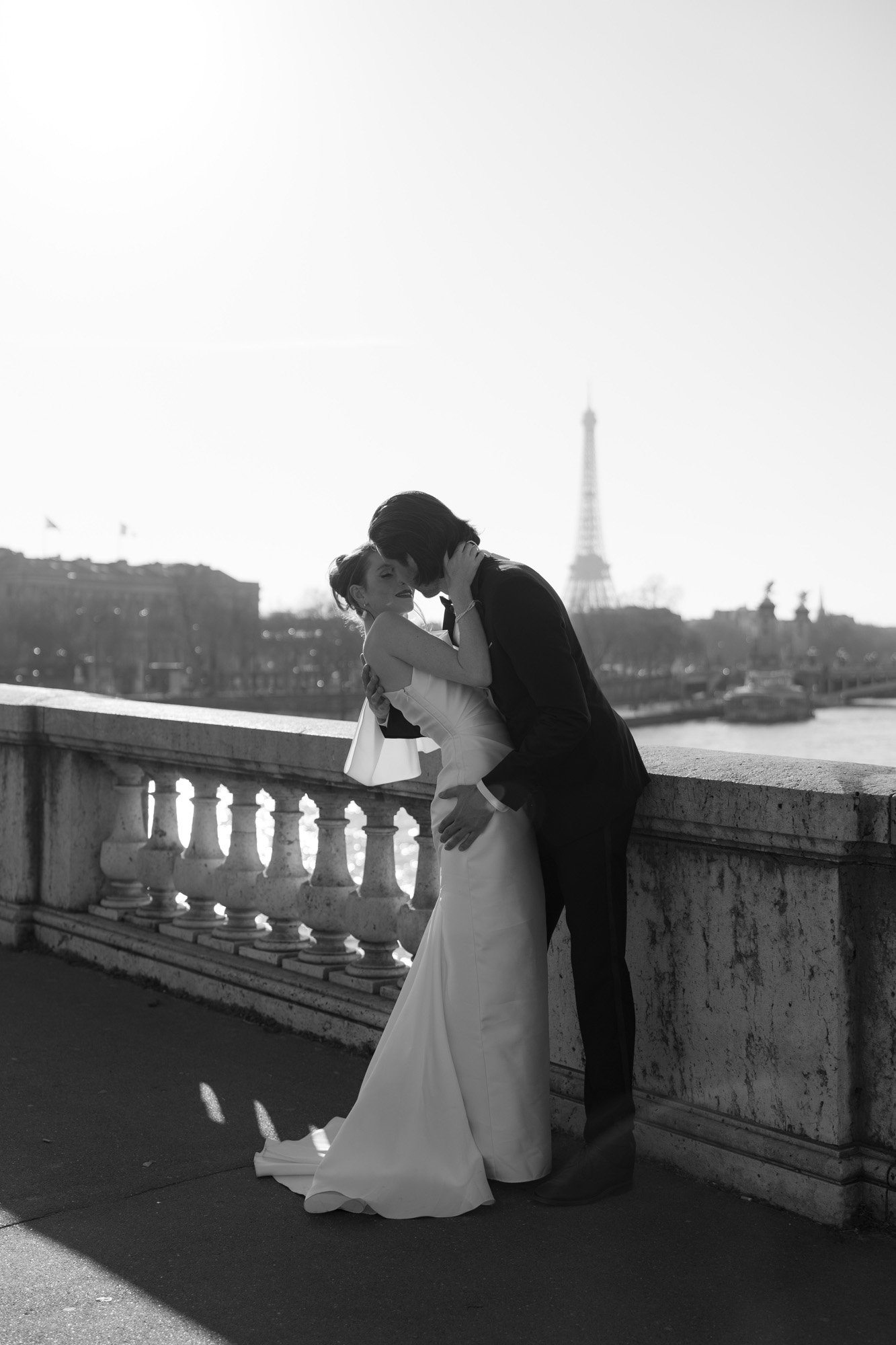 A couple in wedding attire kisses on a stone bridge with the Eiffel Tower visible in the background on a sunny day.