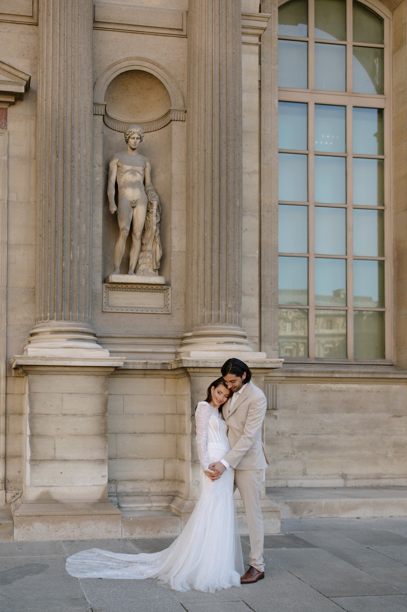 A bride and groom stand embracing in front of a stone building with tall columns, large windows, and a classical statue in the background. Paris city elopement photographer.