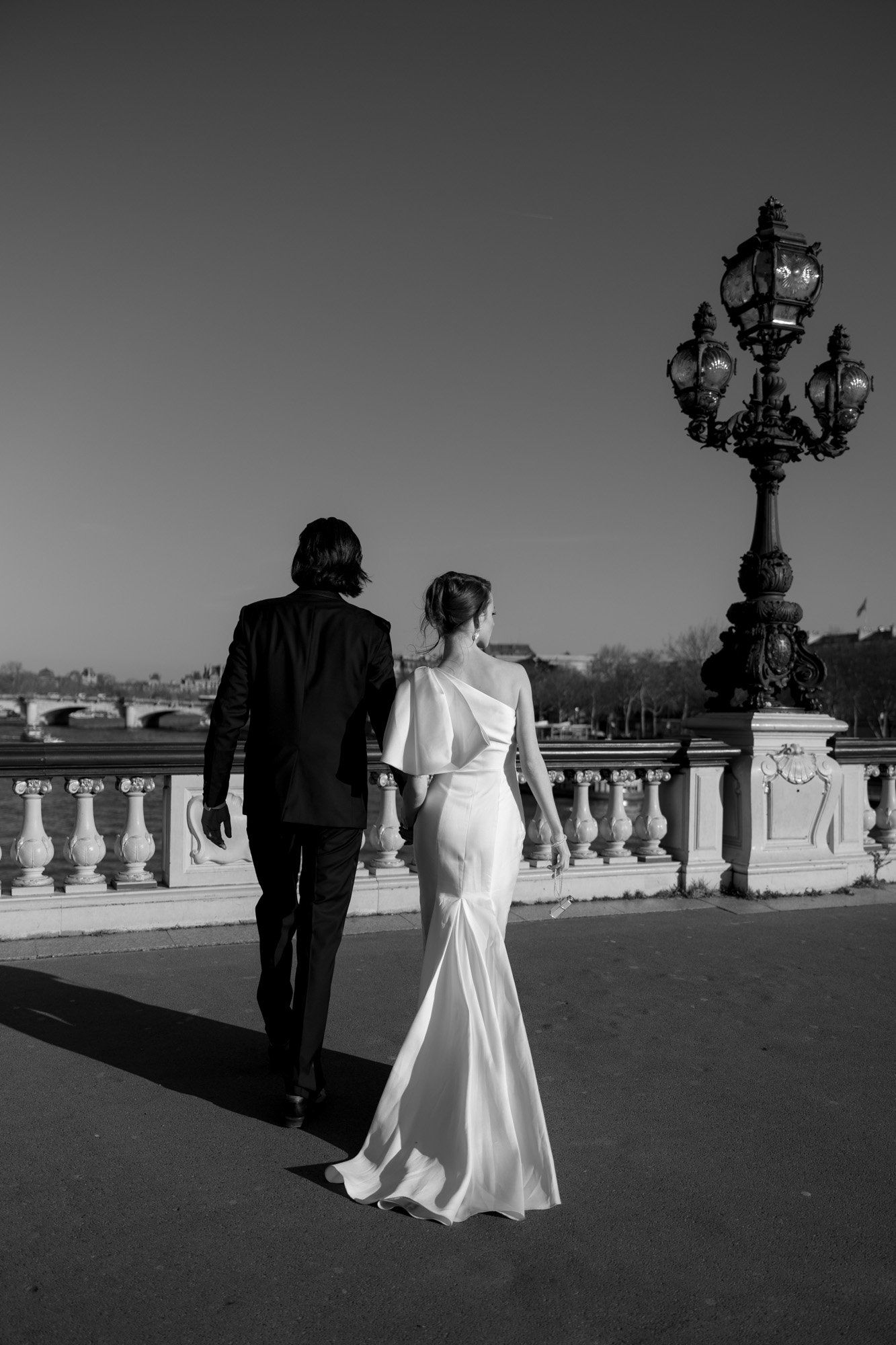 A couple dressed in formal attire, with the woman in a white gown, walk side by side on a bridge with ornate lamp posts under a clear sky.