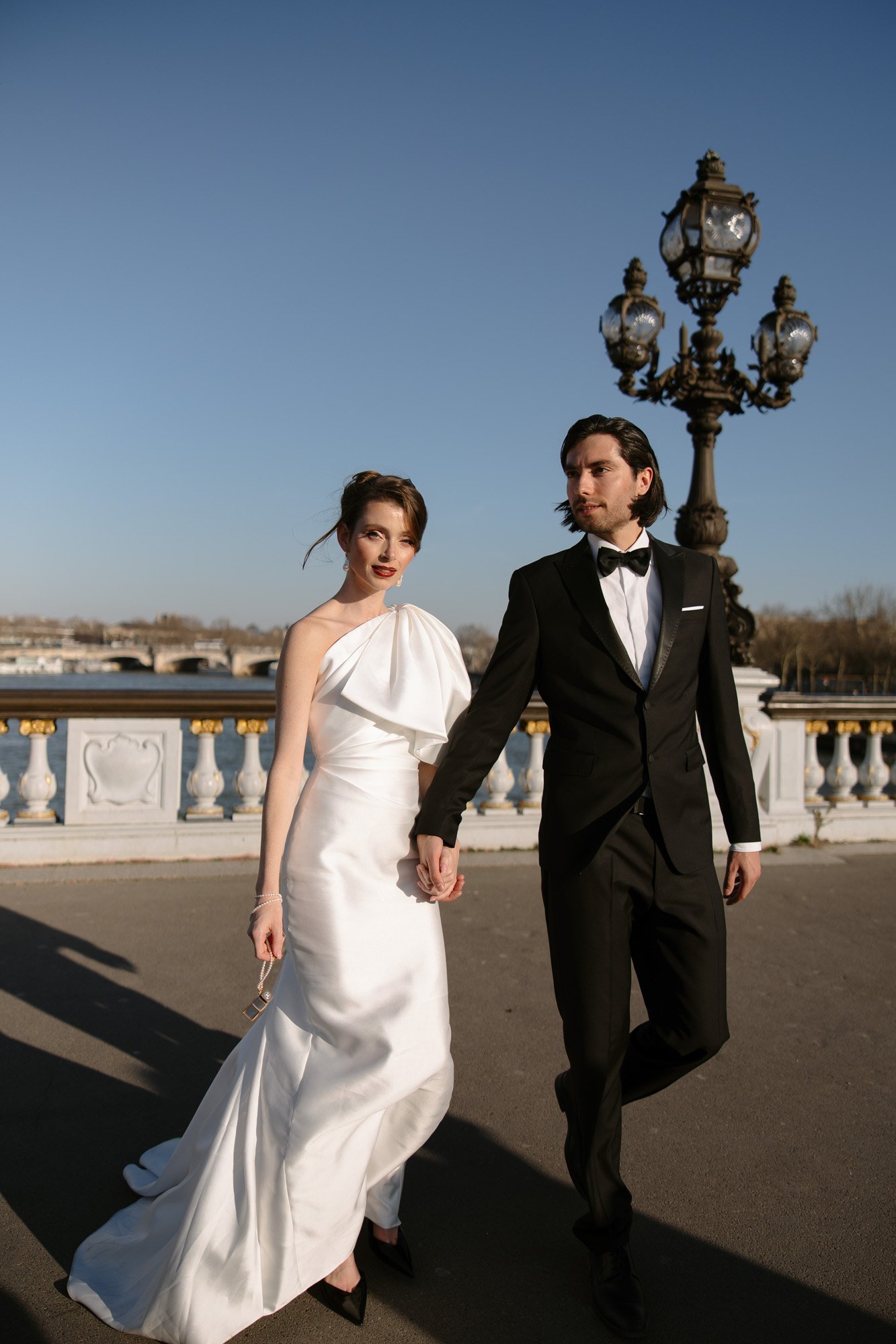 A man in a black tuxedo and a woman in a white gown hold hands while standing outdoors near an ornate street lamp and a bridge. Paris city elopement photographer.