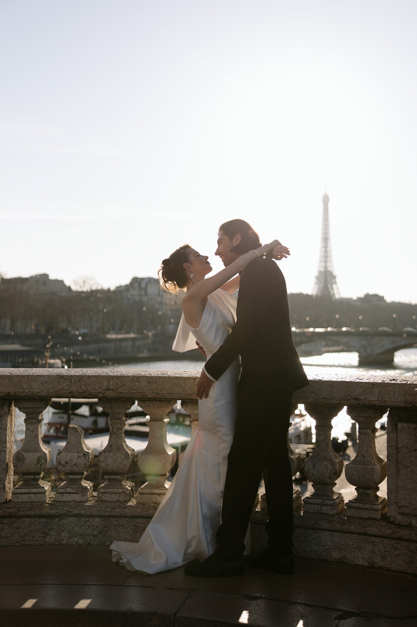 A couple in formal attire embraces on a stone terrace with the Eiffel Tower and a river in the background at sunset. Paris wedding photographer.