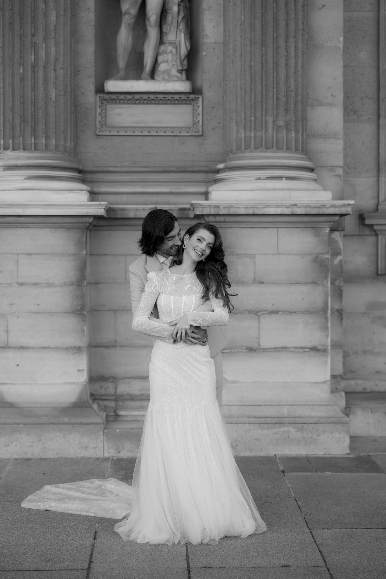 A couple in wedding attire embraces and smiles in front of a stone building with columns and a statue above them.