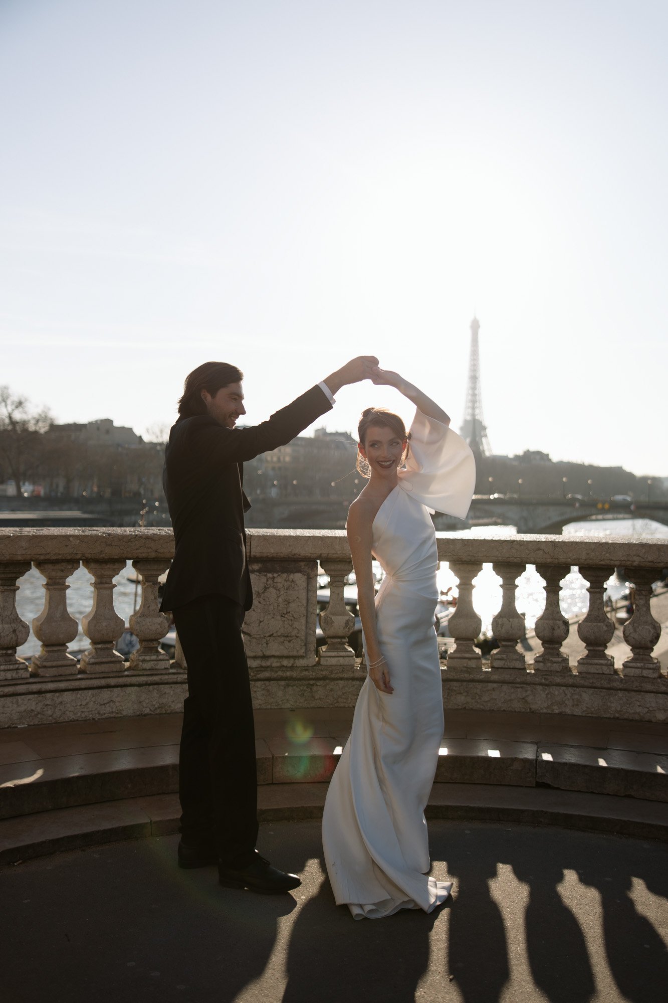 A couple dressed in formal attire dance on a stone terrace with the Eiffel Tower and river in the background during sunset.