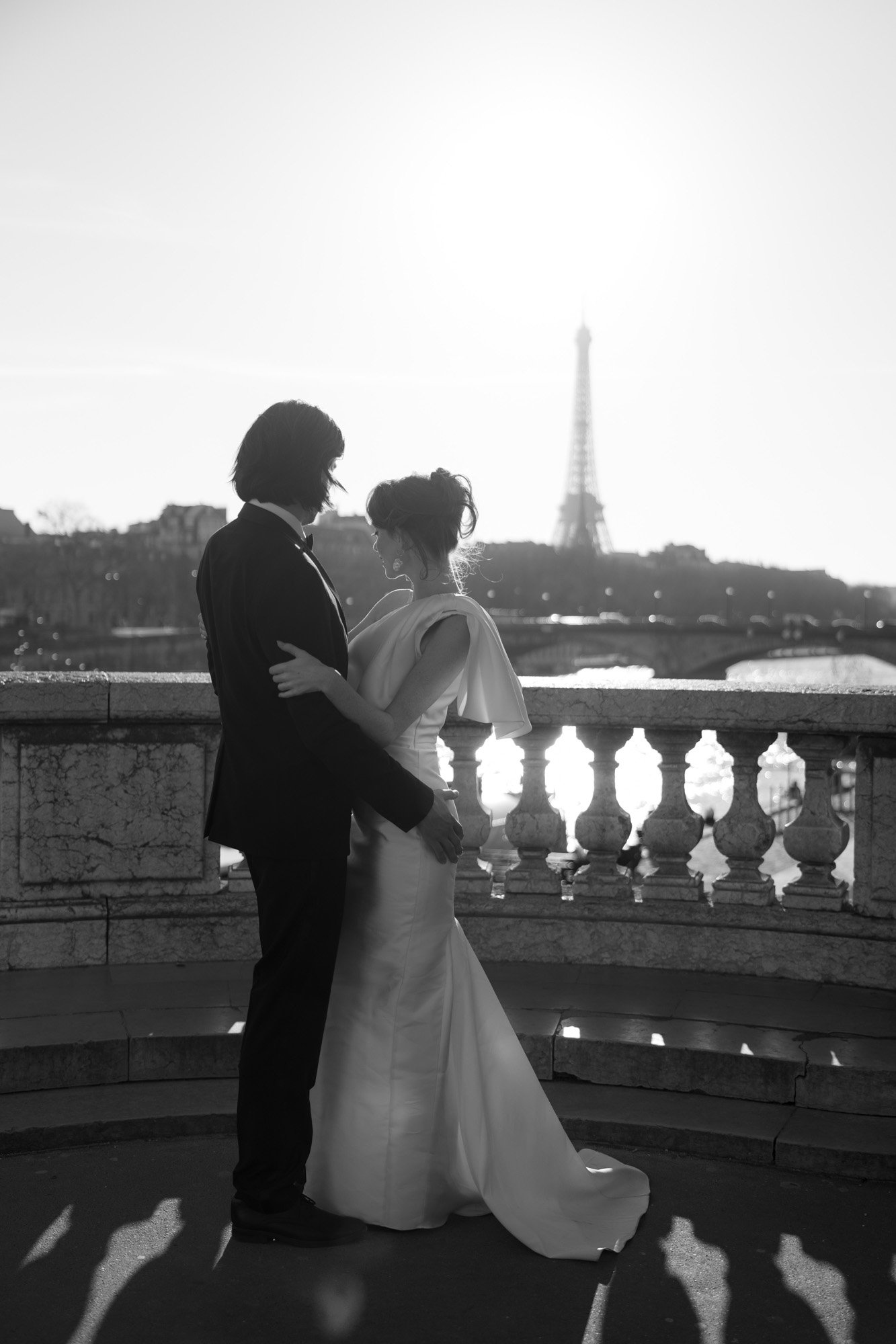 A couple in formal attire stands close together on a terrace, with the Eiffel Tower and a bright sun visible in the background.