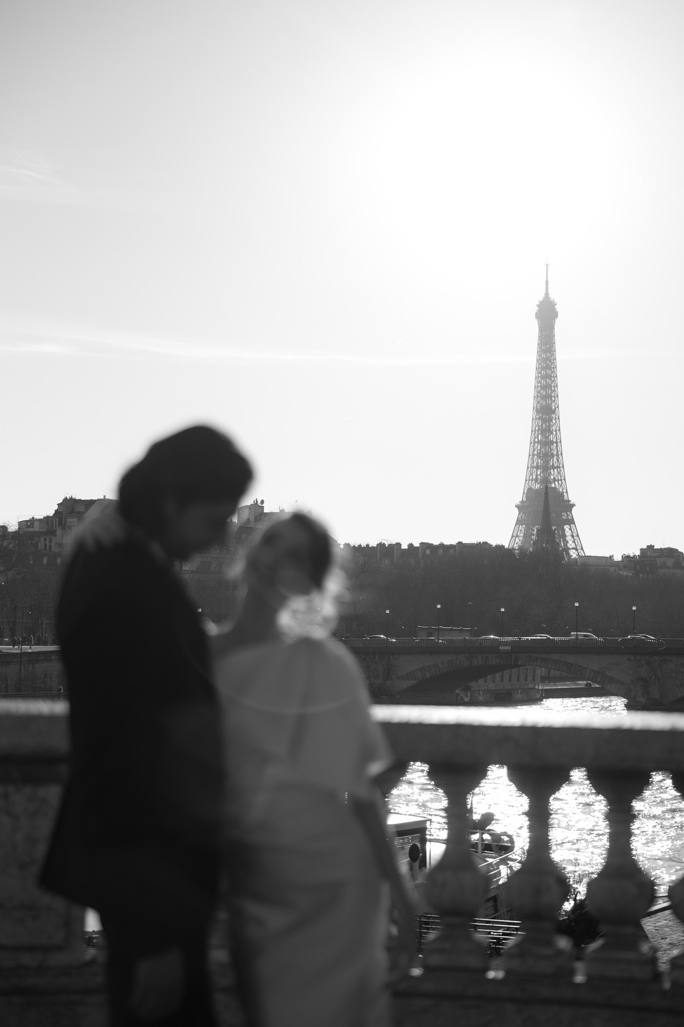 A couple stands close together on a bridge with the Eiffel Tower in the background, captured in black and white with both people out of focus.