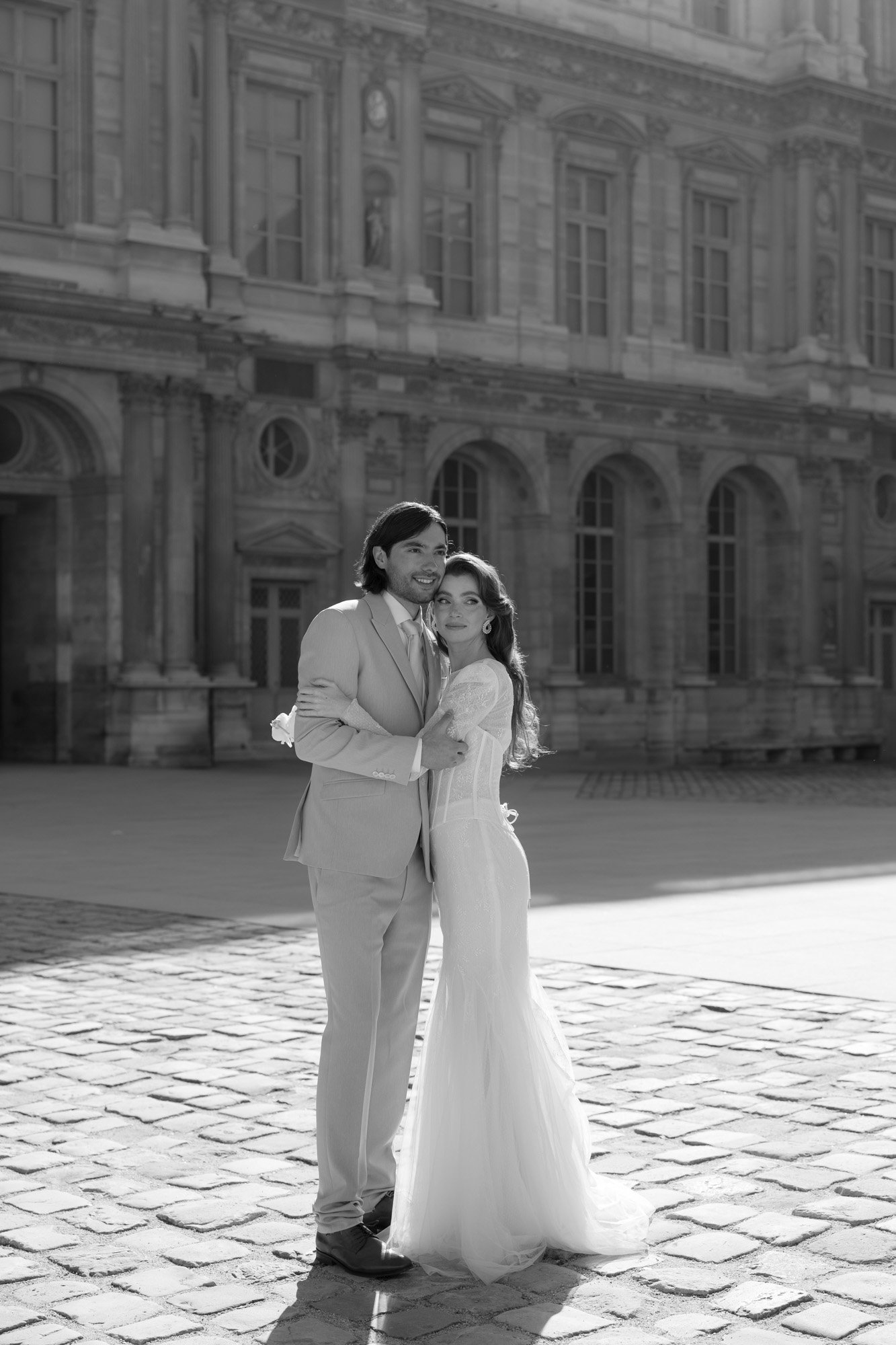 A couple dressed in formal wedding attire stands together on a cobblestone courtyard in front of a historic building. Paris city elopement photographer.