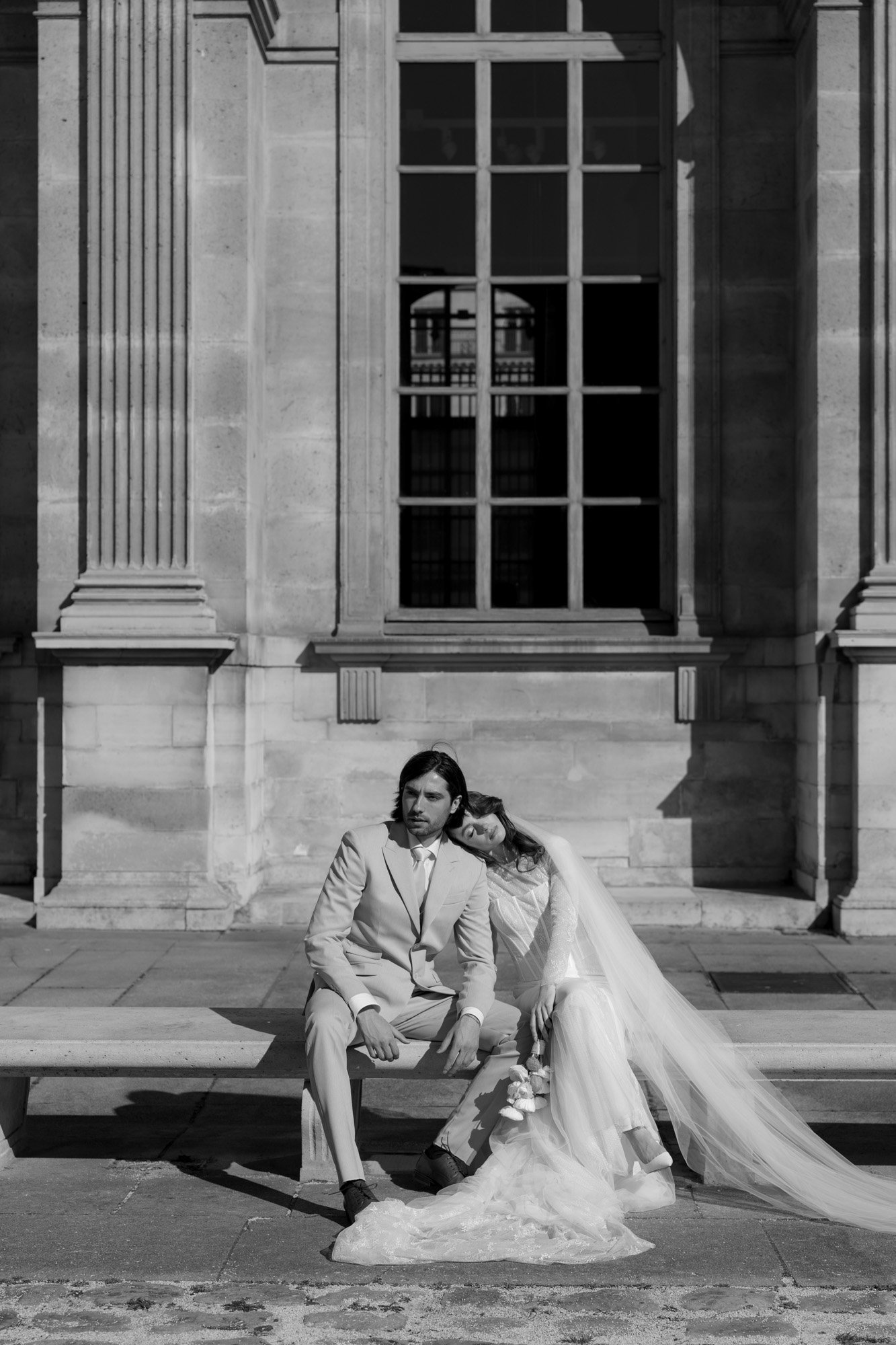 A bride in a wedding dress leans her head on the grooms shoulder as they sit on a bench outside a building with large windows and columns. Paris city elopement photographer.