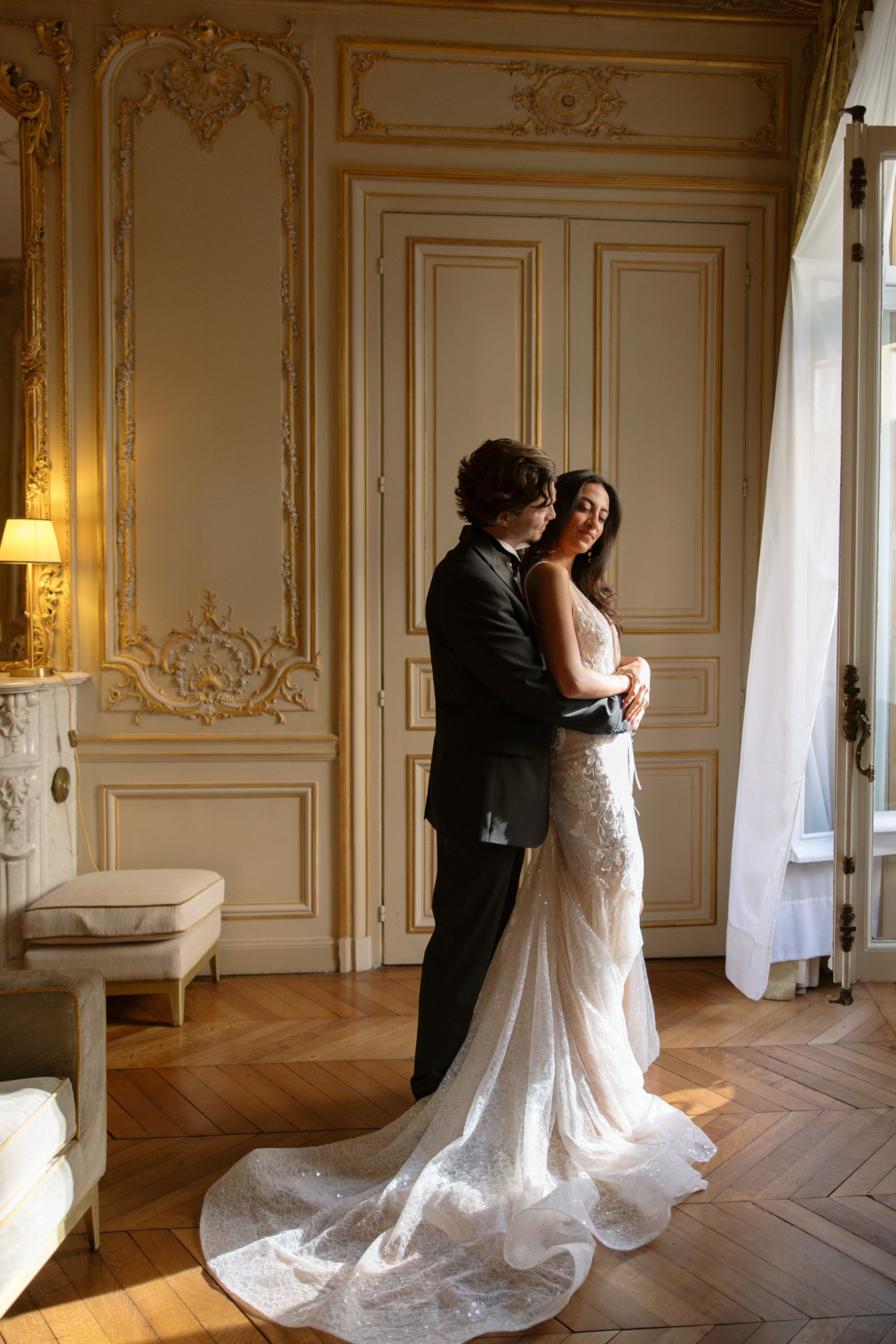 A couple in formal wedding attire stands embracing near a window in an ornate, sunlit room with gold-trimmed walls and wooden floors. Hotel Alfred Sommier Elopement.