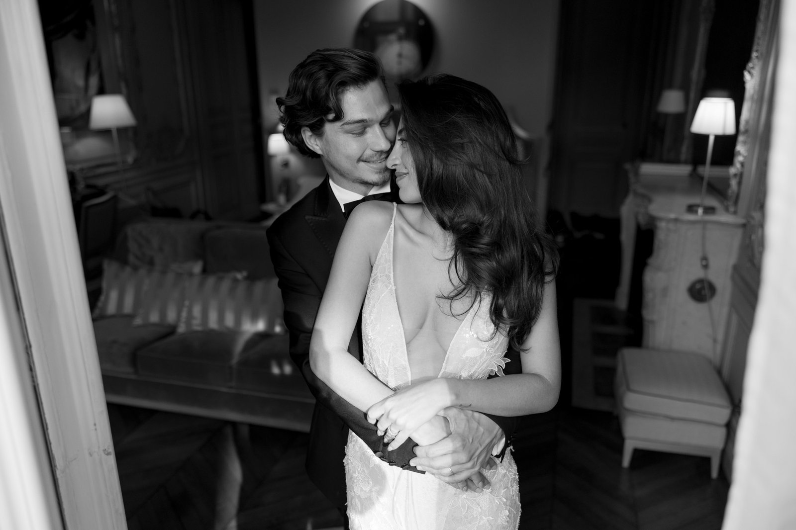 A man in a suit embraces a woman in a white dress from behind in an elegantly decorated room. Both appear happy and affectionate. Hotel Alfred Sommier Elopement.