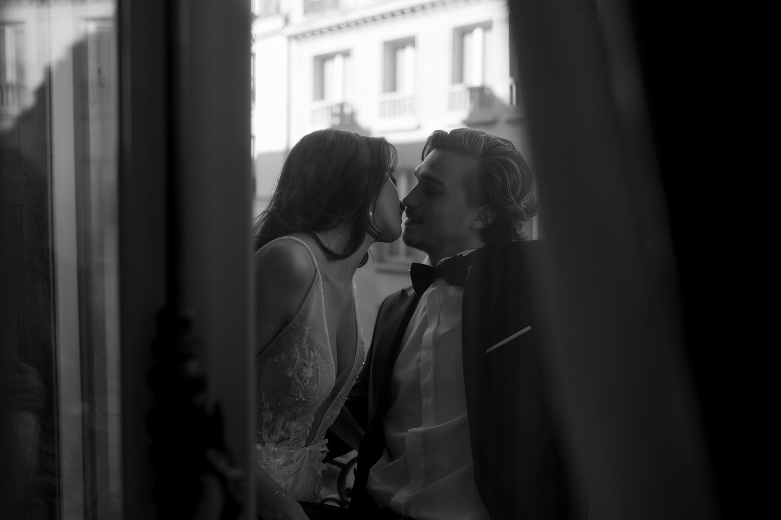 A woman in a dress leans in to kiss a man in a suit and bow tie, sitting closely together by a window; the scene is in black and white. Hotel Alfred Sommier Elopement.