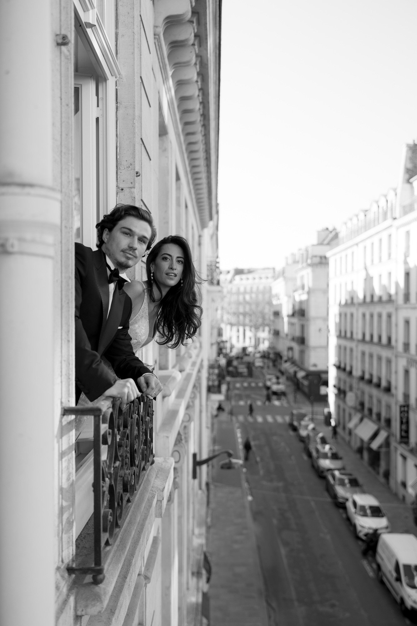 A man and a woman in formal attire lean out of a window on a city street, looking into the distance; several buildings and parked cars are visible below. Hotel Alfred Sommier Elopement.