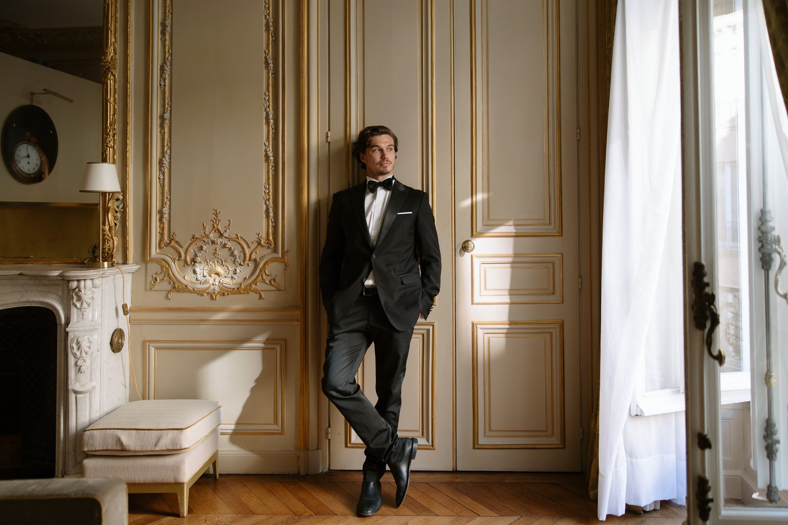 A man in a black tuxedo stands with crossed legs against ornate cream-colored paneled doors in a classic, elegant room. Hotel Alfred Sommier Elopement.