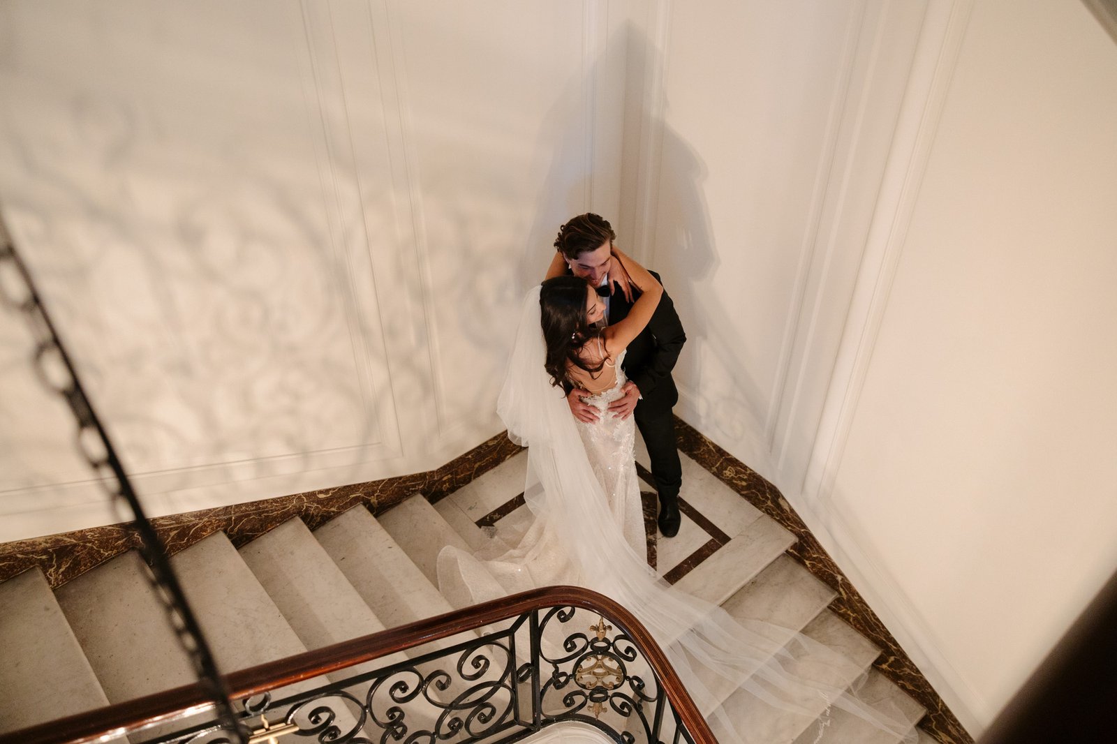 A bride and groom embrace on a marble staircase, with the bride’s veil flowing down the steps in an elegant indoor setting. Hotel Alfred Sommier Elopement.