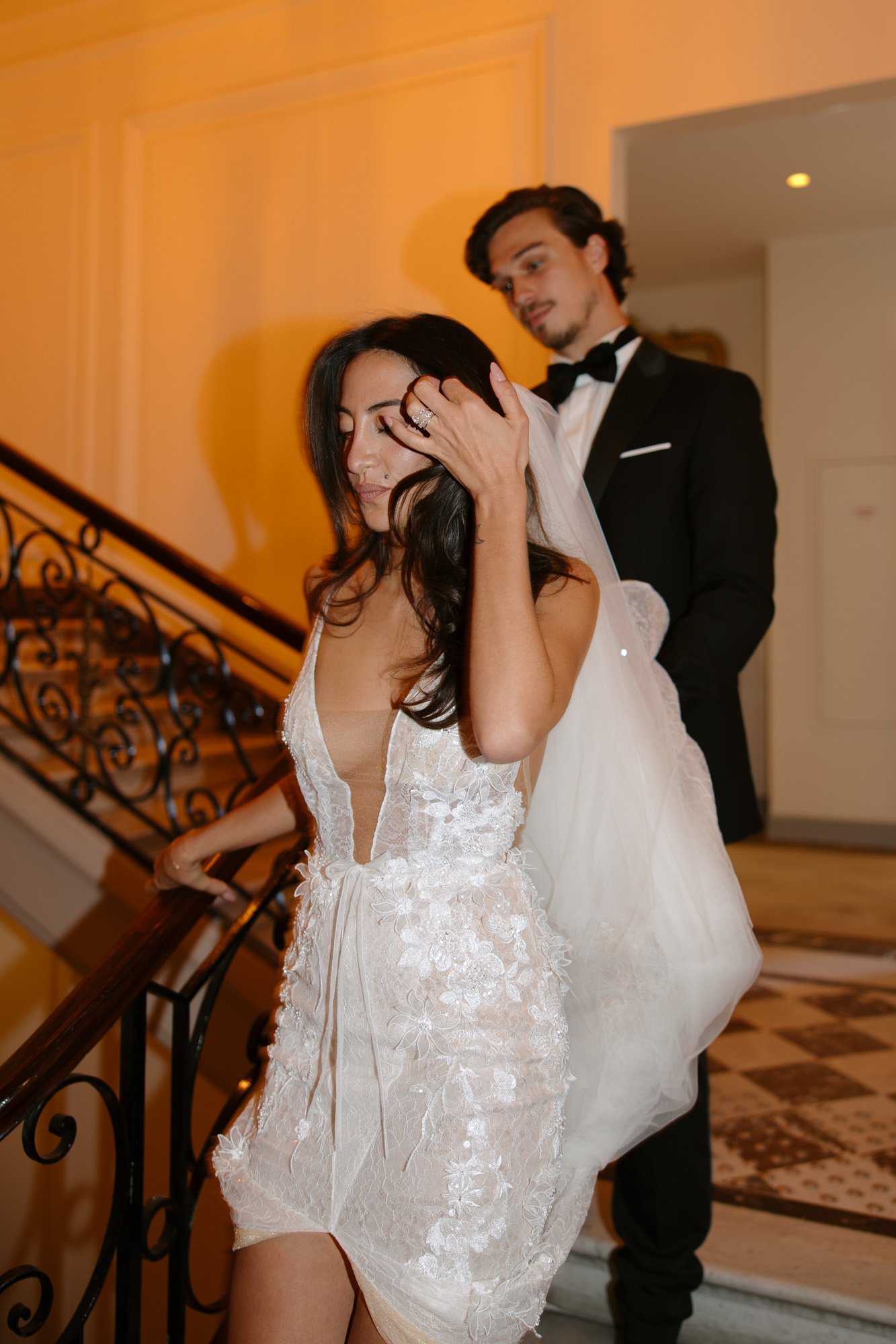 A woman in a white wedding dress descends a staircase, adjusting her hair, while a man in a black suit and bow tie follows behind. Hotel Alfred Sommier Elopement.