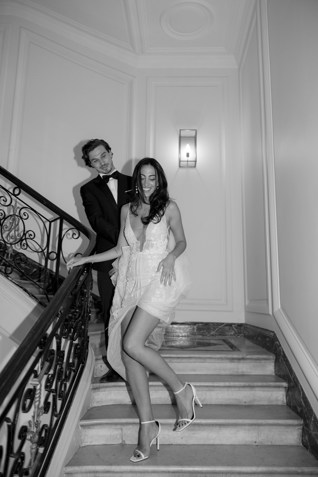 A woman in a light dress and heels descends a staircase while a man in a tuxedo stands behind her. The setting appears formal, with ornate railings and paneled walls. Hotel Alfred Sommier Elopement.