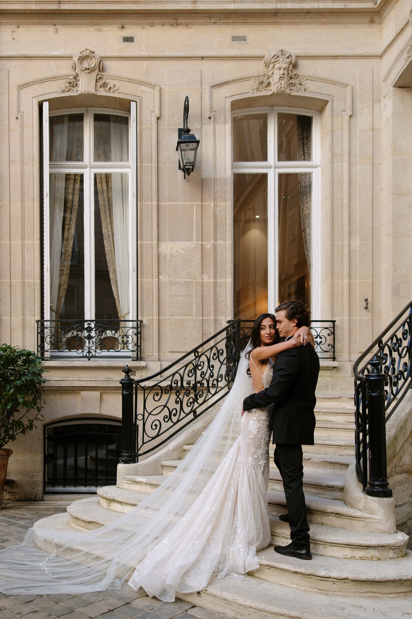 A bride and groom stand on a curved stone staircase outside a grand building, embracing and looking at the camera. The bride wears a long, white gown with a train. Hotel Alfred Sommier Elopement.