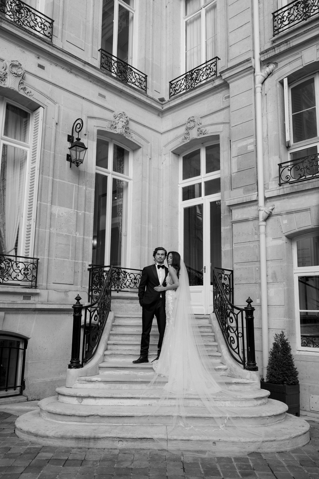 A bride and groom stand on a stone staircase outside a large, elegant building, dressed in formal wedding attire. Hotel Alfred Sommier Elopement.